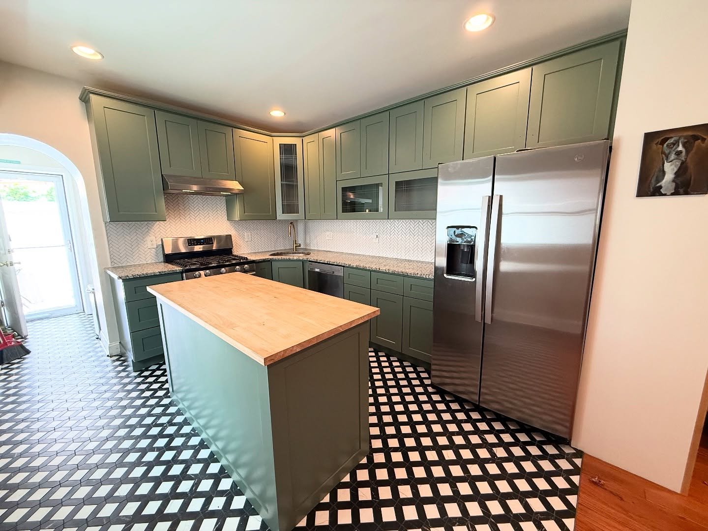 Full view of sage green Rosepine kitchen with butcher block island, black and white checkered floor, stainless side by side fridge, and matching sage uppers and lowers.
