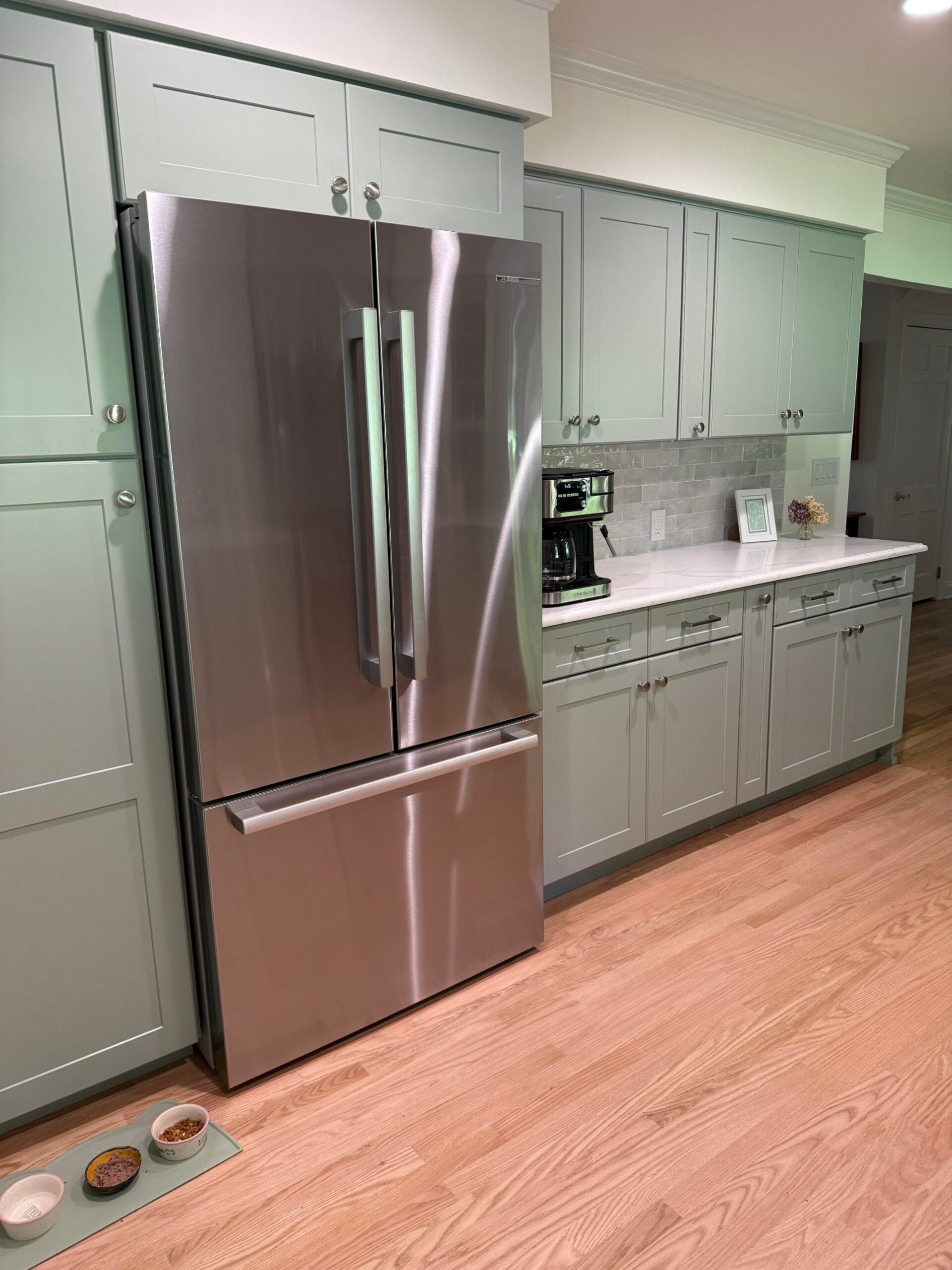 Full-height sage green shaker pantry tower on the left, a stainless French-door fridge, and a run of sage shaker base and wall cabinets with a coffee station on the right.
