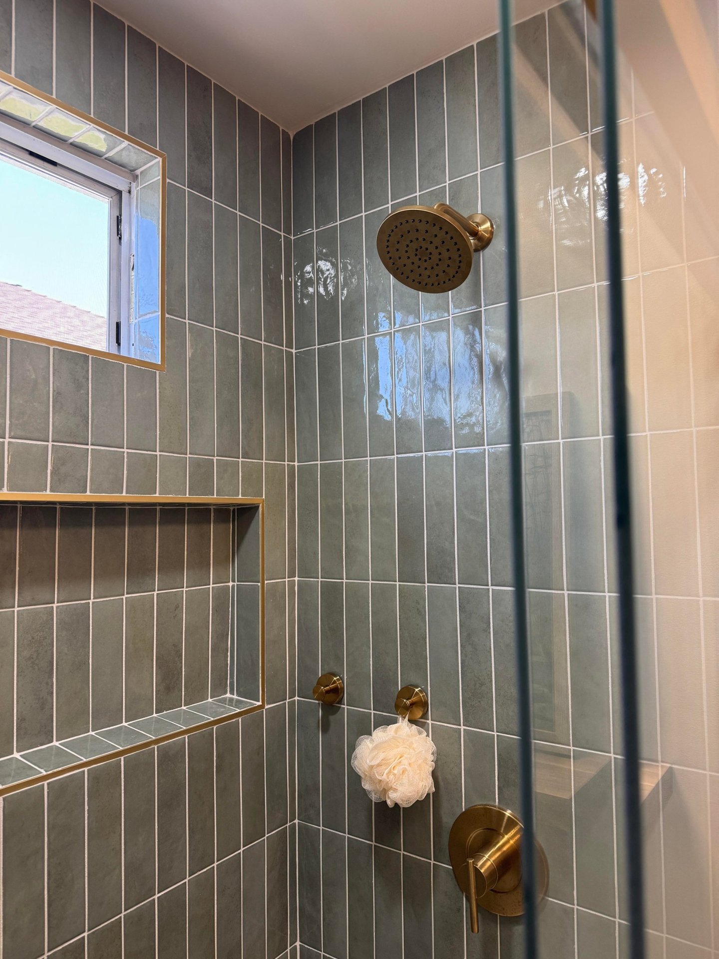 Close up of the sage green vertical tile shower showing a brass trimmed wall niche, brass round shower head, brass valve, and a small frosted window with brass casing.