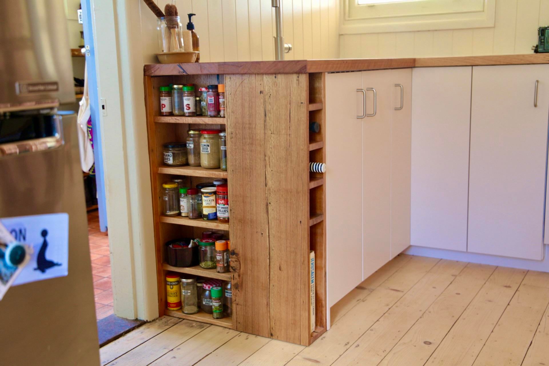 Slim open wood spice rack filled with spice jars, built into the end of a white kitchen cabinet run, with a narrow side slot for bottles and a book.