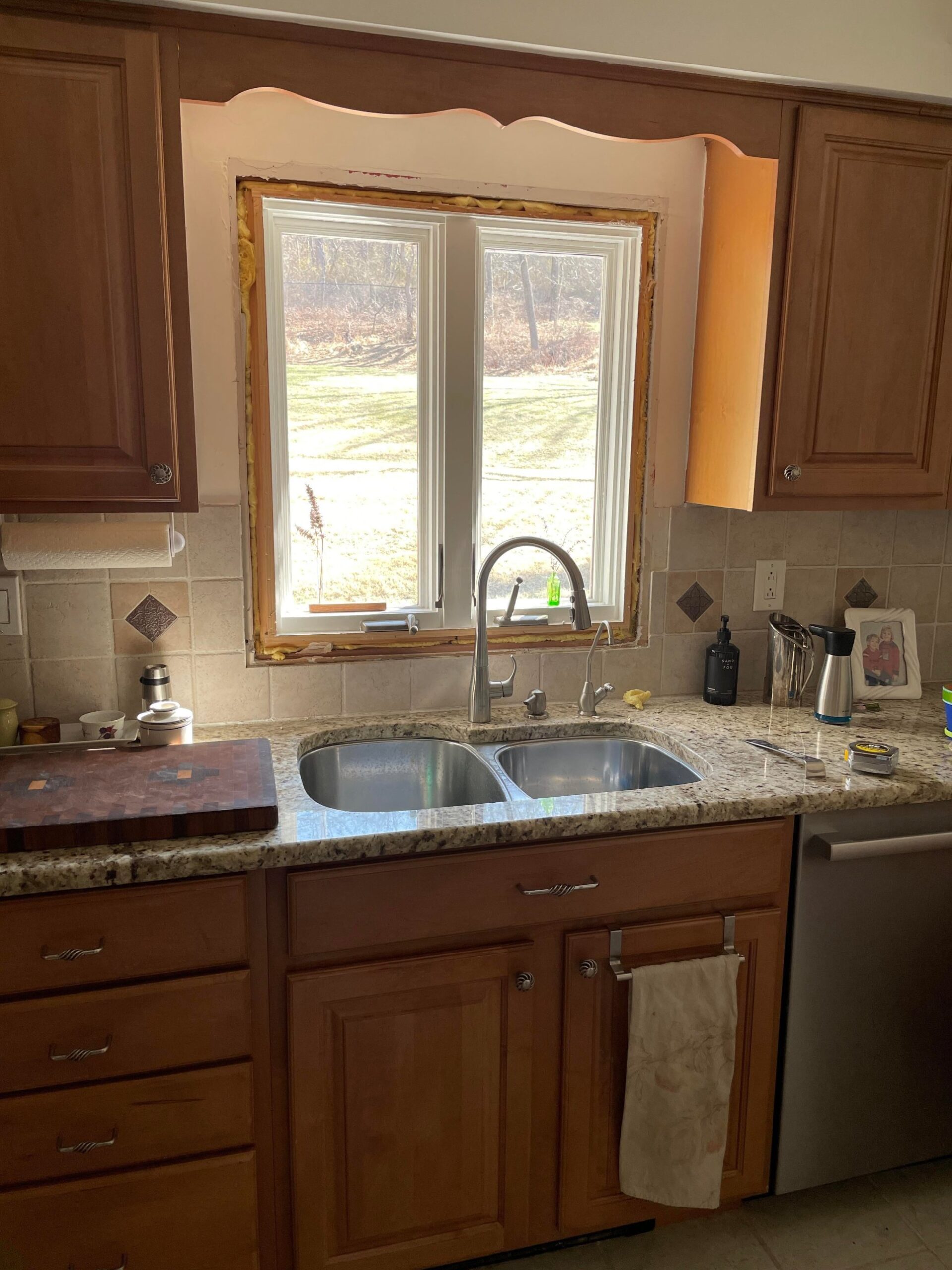 Honey oak kitchen sink wall with a scalloped wood valance above the window, old granite counter, and a stainless double-basin sink.