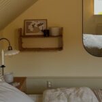Cozy Scandinavian attic bedroom with a sloped white beadboard ceiling, warm butter yellow walls, a bed dressed in white linen, a brown gingham duvet, and a matching pillow, a small wood nightstand with a brass gooseneck wall sconce and a lit taper candle, a woven rattan wall shelf holding art and a candle, and a long oval black-framed mirror reflecting the window’s warm afternoon light.