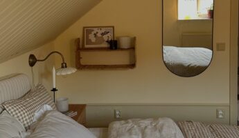 Cozy Scandinavian attic bedroom with a sloped white beadboard ceiling, warm butter yellow walls, a bed dressed in white linen, a brown gingham duvet, and a matching pillow, a small wood nightstand with a brass gooseneck wall sconce and a lit taper candle, a woven rattan wall shelf holding art and a candle, and a long oval black-framed mirror reflecting the window’s warm afternoon light.