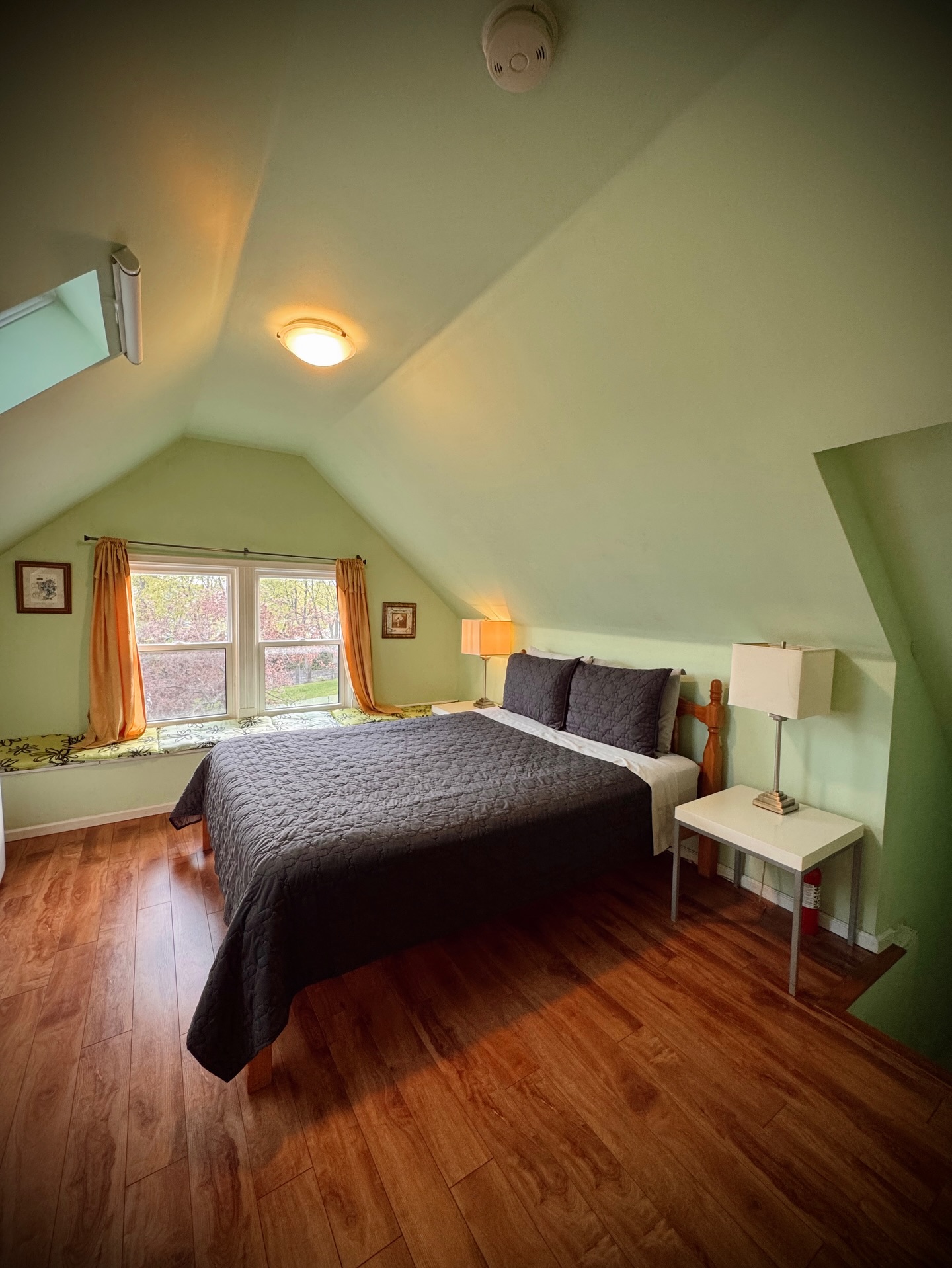 Simple attic bedroom with pale mint green walls and sloped ceiling, dark gray quilted bedding on a traditional spindle bed, orange curtains, window seat with yellow and green cushion, and warm wood plank floors