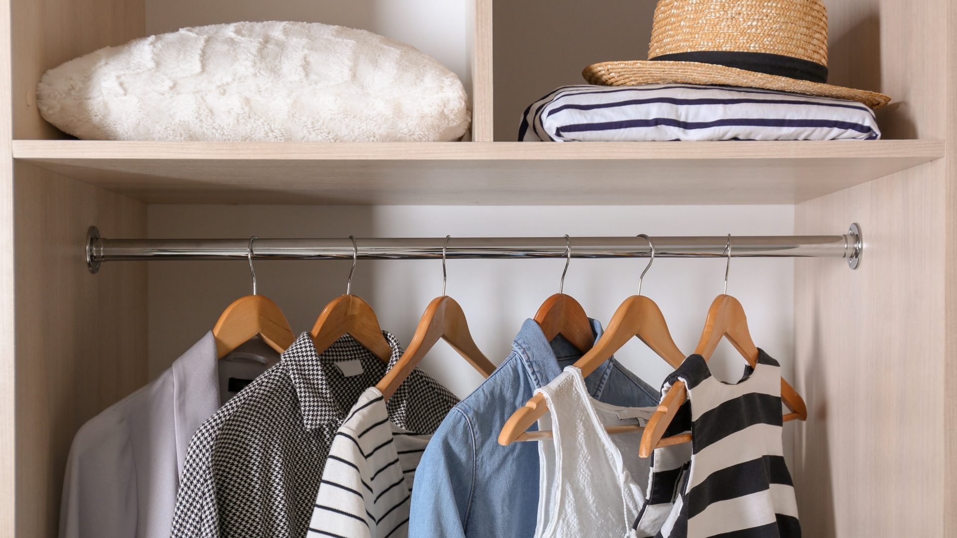 Open wardrobe with a single chrome rod holding shirts and tops on wooden hangers, including a houndstooth shirt, a denim shirt and striped tops, under a shelf with a fluffy white pillow, a straw hat and folded striped fabric.