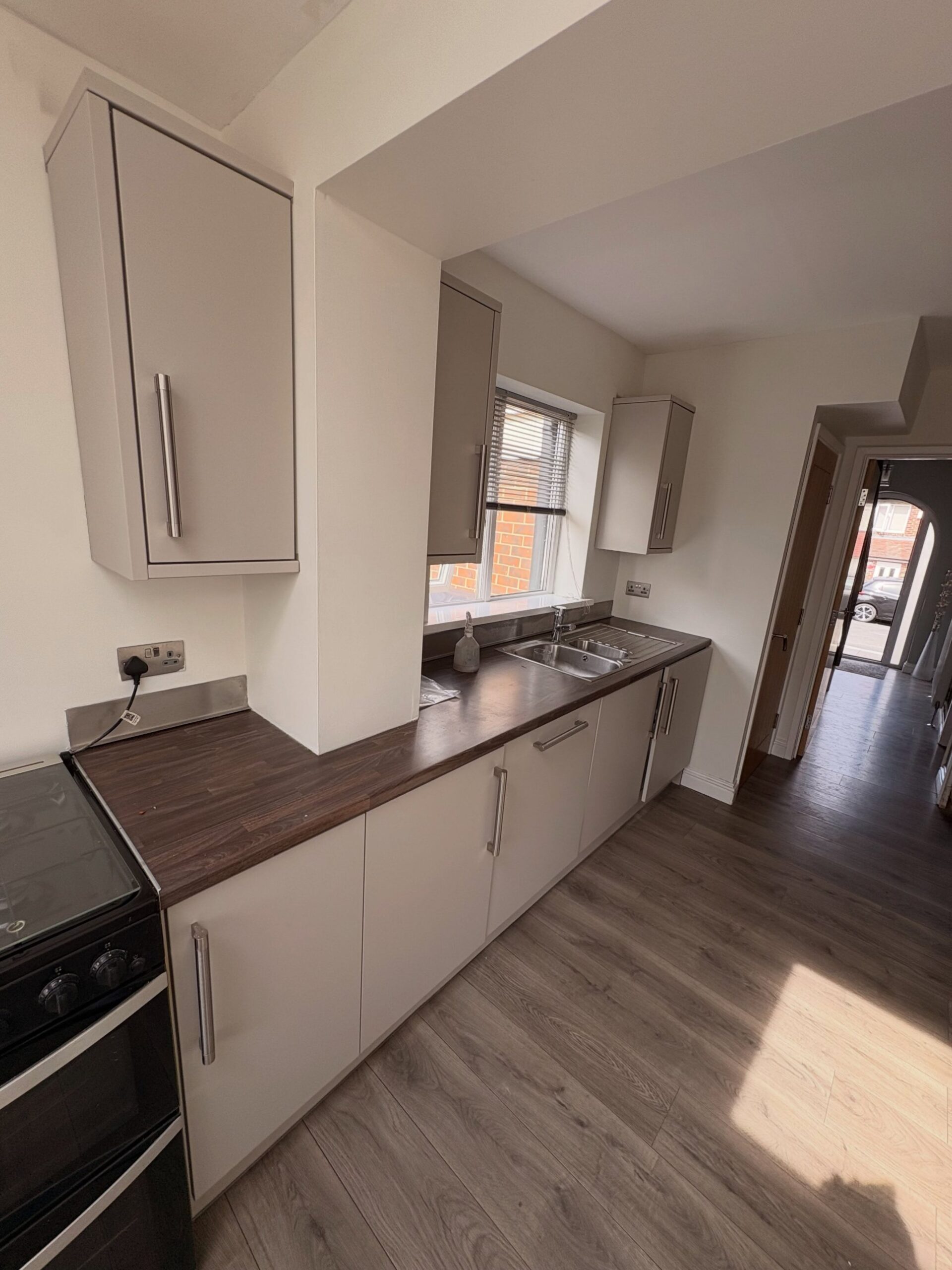 Sink and window end of the kitchen freshly sprayed in Purbeck Stone, dark walnut counter, stainless steel sink under the window, hallway visible to the right leading to the front door.