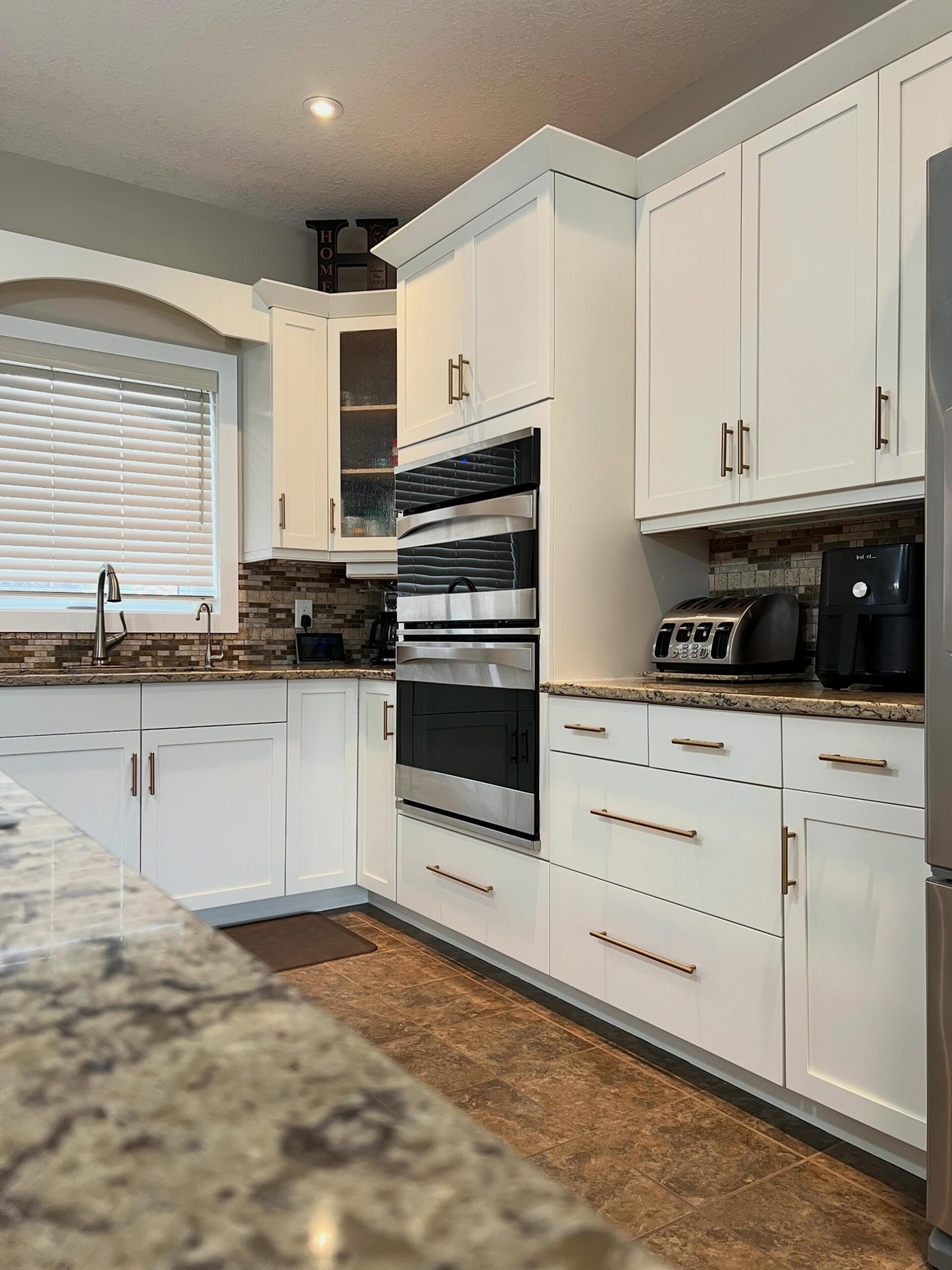 Corner angle of the Soft Chamois kitchen showing the sink window on the left, the double wall oven centered, upper and lower shaker cabinets with champagne bronze pulls, speckled granite counters, mosaic backsplash, and the island counter in the foreground.