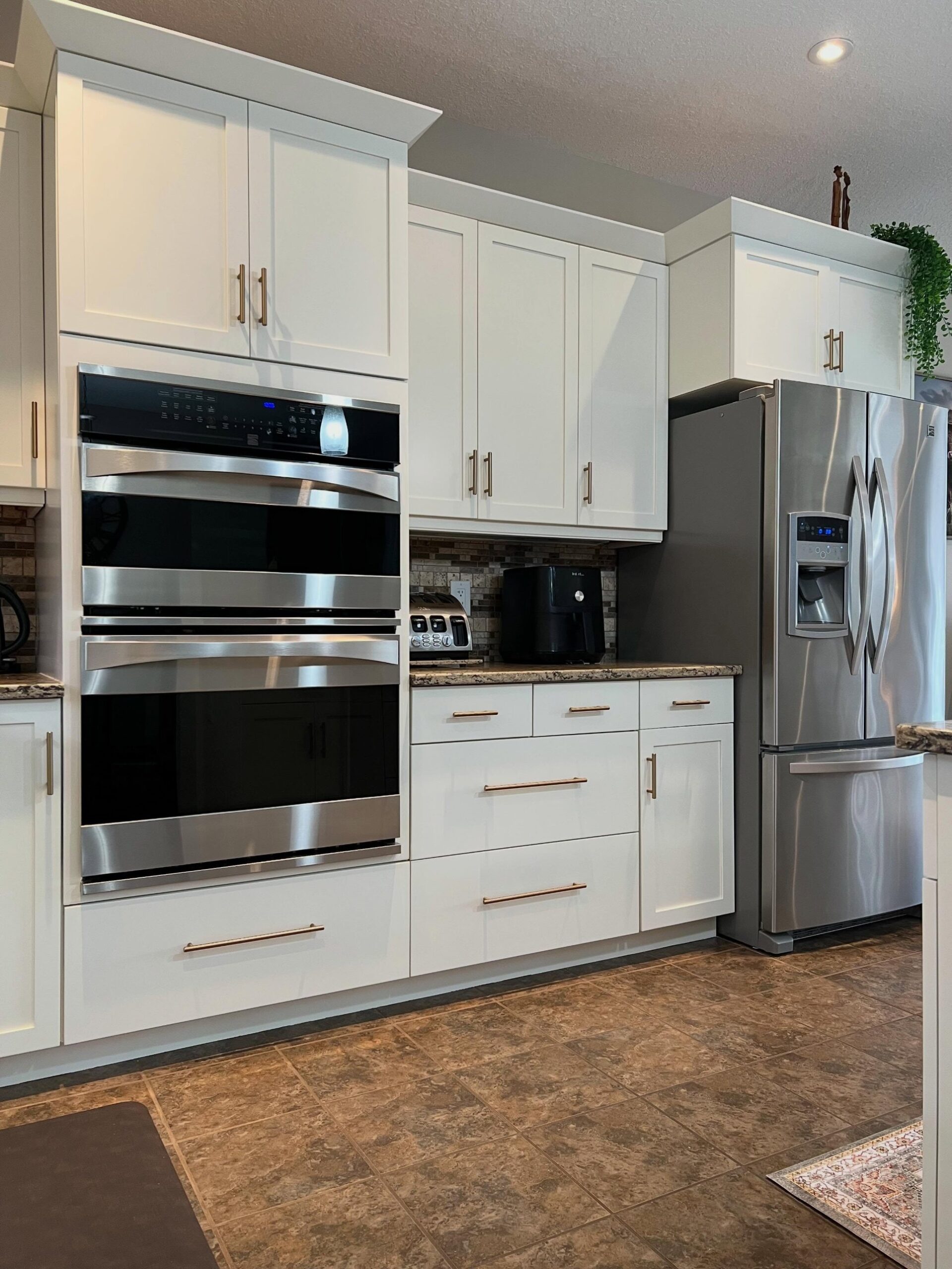 Close view of the double stainless wall oven set into Soft Chamois cabinets with long champagne bronze bar pulls on the drawers below, the mosaic backsplash and granite counter visible, and the brown tile floor at the base.