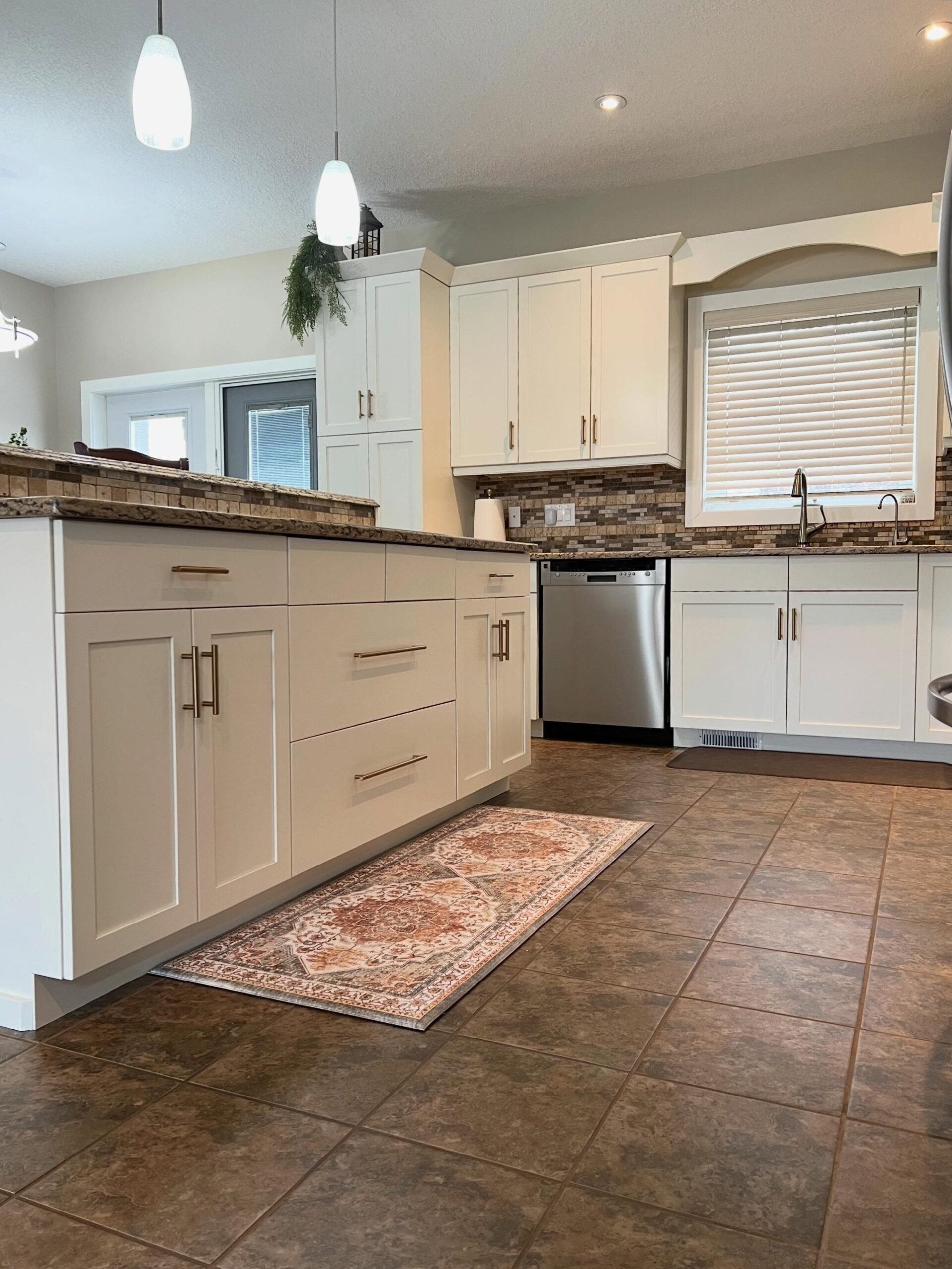 Side angle of the painted island back in Soft Chamois with long champagne bronze bar pulls, a terracotta-and-cream Persian-style runner rug on the brown floor tile, and the sink wall visible in the background.