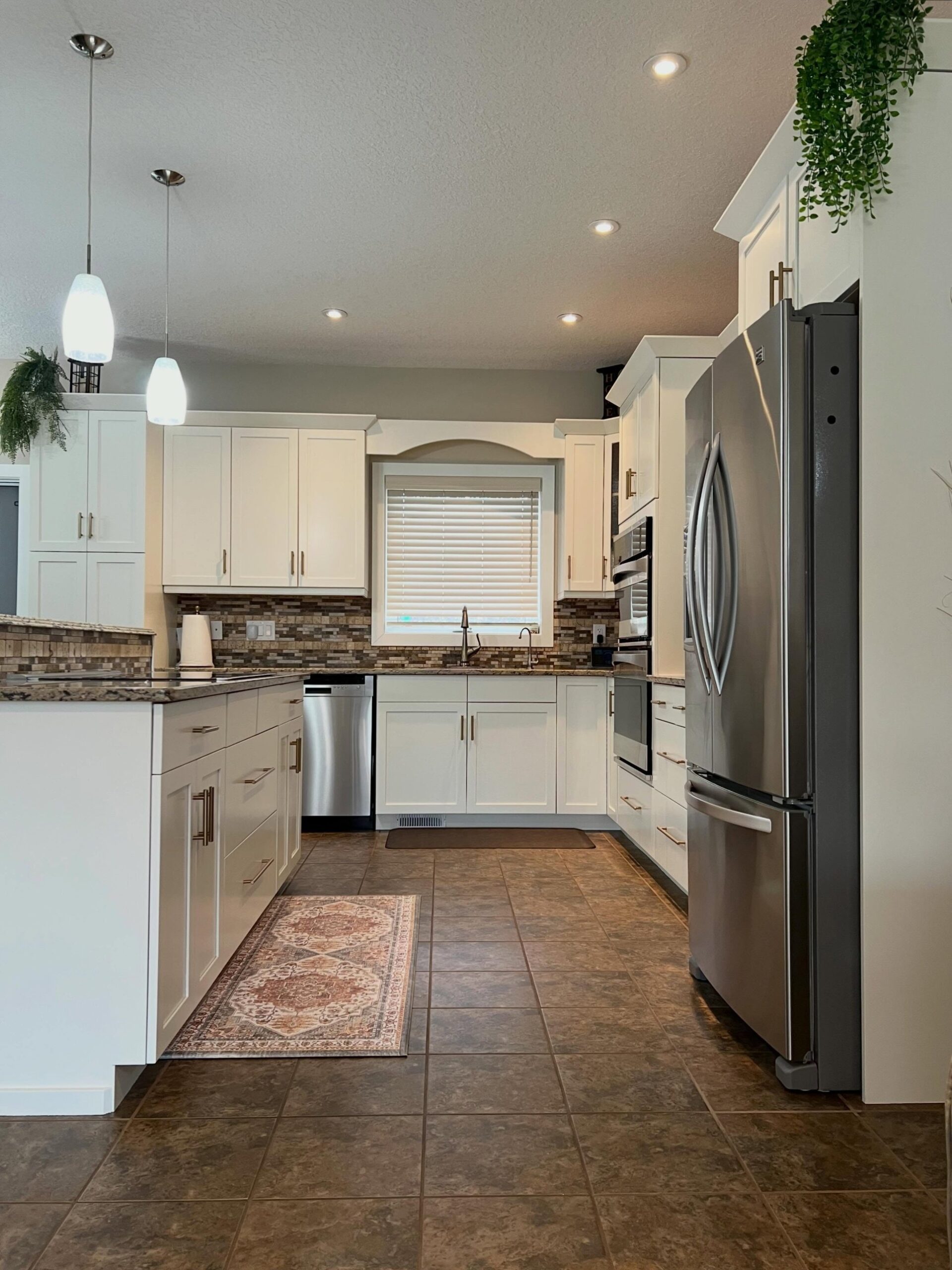 Head-on view of the same kitchen after being painted Benjamin Moore Soft Chamois, with champagne bronze pulls, a Persian-style runner rug on the brown floor tile, trailing greenery over the fridge, and the same arched valance over the sink window.