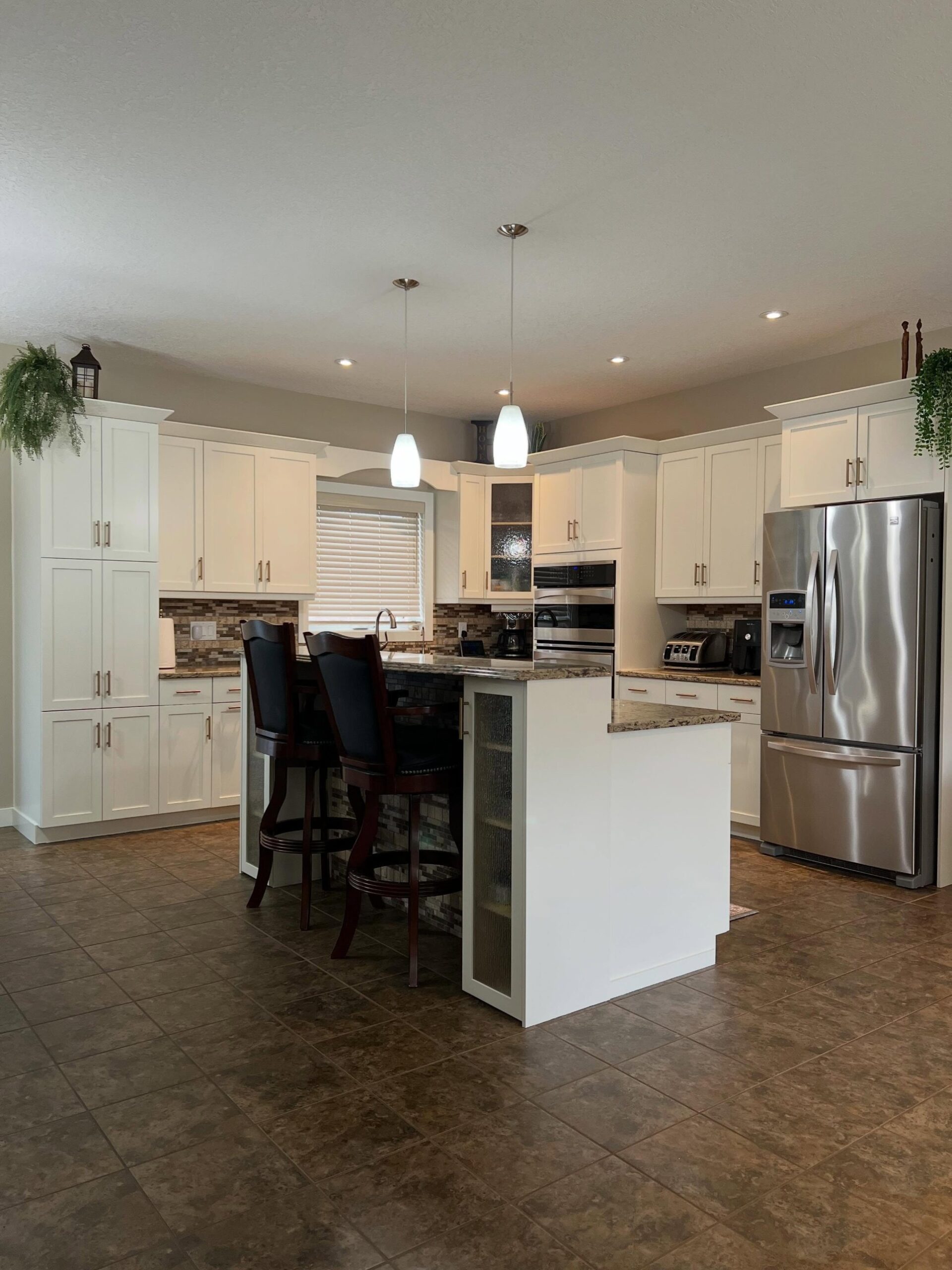 Wide view of a white kitchen with Benjamin Moore Soft Chamois shaker cabinets, champagne bronze pulls, gold-and-brown speckled granite counters, a tumbled stone mosaic backsplash, brown ceramic floor tile, two glass pendant lights, dark wood bar chairs at a white island, and a stainless steel French door fridge.