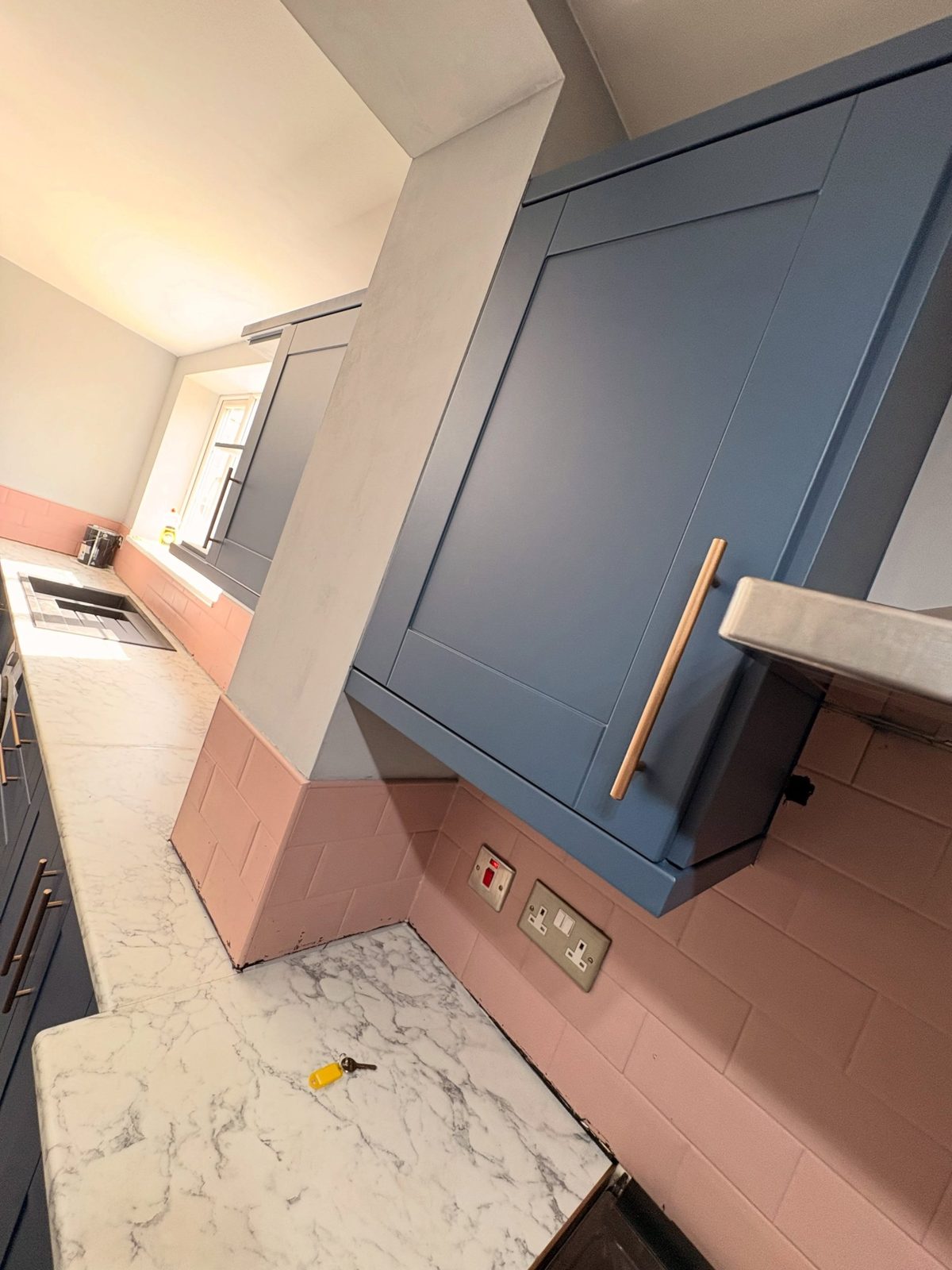 Close view of a tall Stiffkey Blue cabinet with a copper handle sitting flush against a dusty pink subway tile wall, with a white marble counter running past.