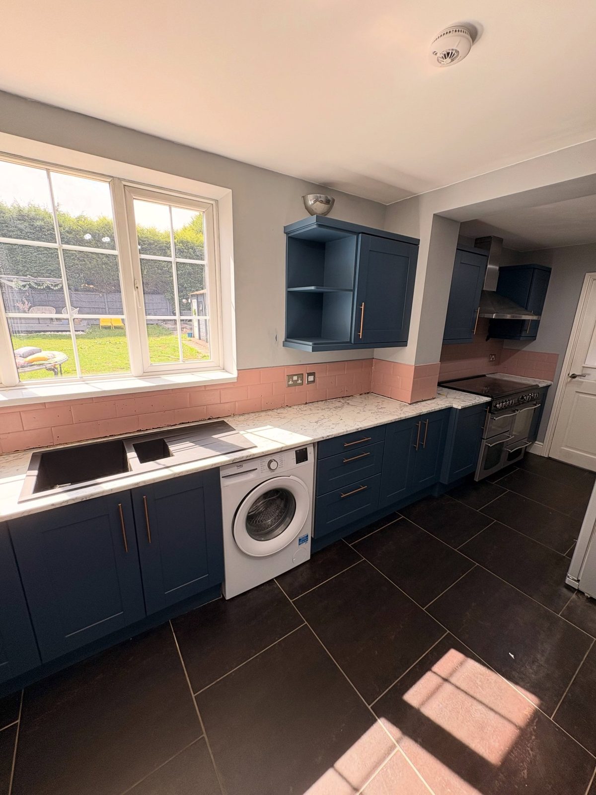 Full wide view of the finished Stiffkey Blue kitchen with copper handles, white marble counter, pink subway tile band, built-in Beko washer, black range cooker, and a bright window looking onto the garden on the left.