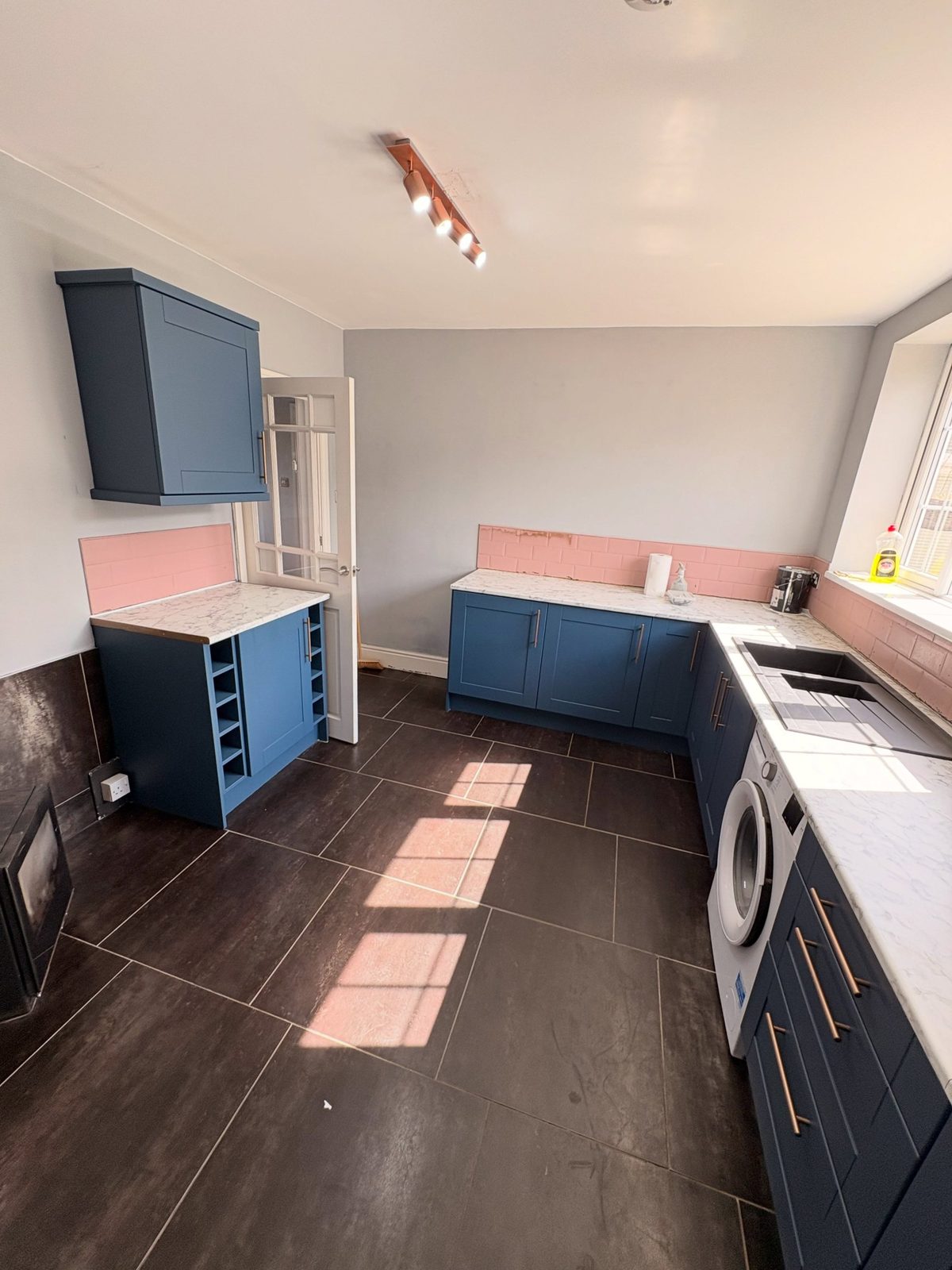 Wide view of the finished Stiffkey Blue kitchen showing a wine rack cabinet on the left, long L-shape run with washer and sink on the right, pink tile band, marble counter, and copper spotlight track overhead.
