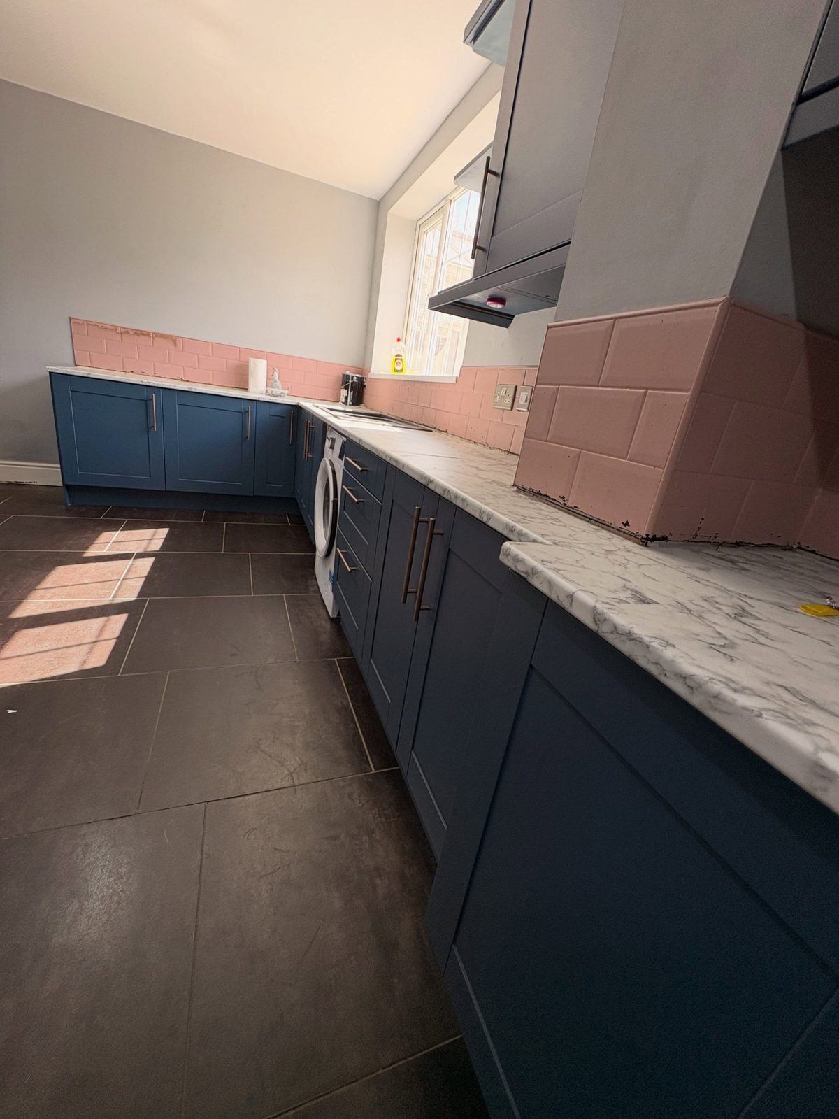 Stiffkey Blue L-shape kitchen cabinets with copper handles, white marble counter, pink subway tile backsplash, and a built-in white washer beside a black sink.