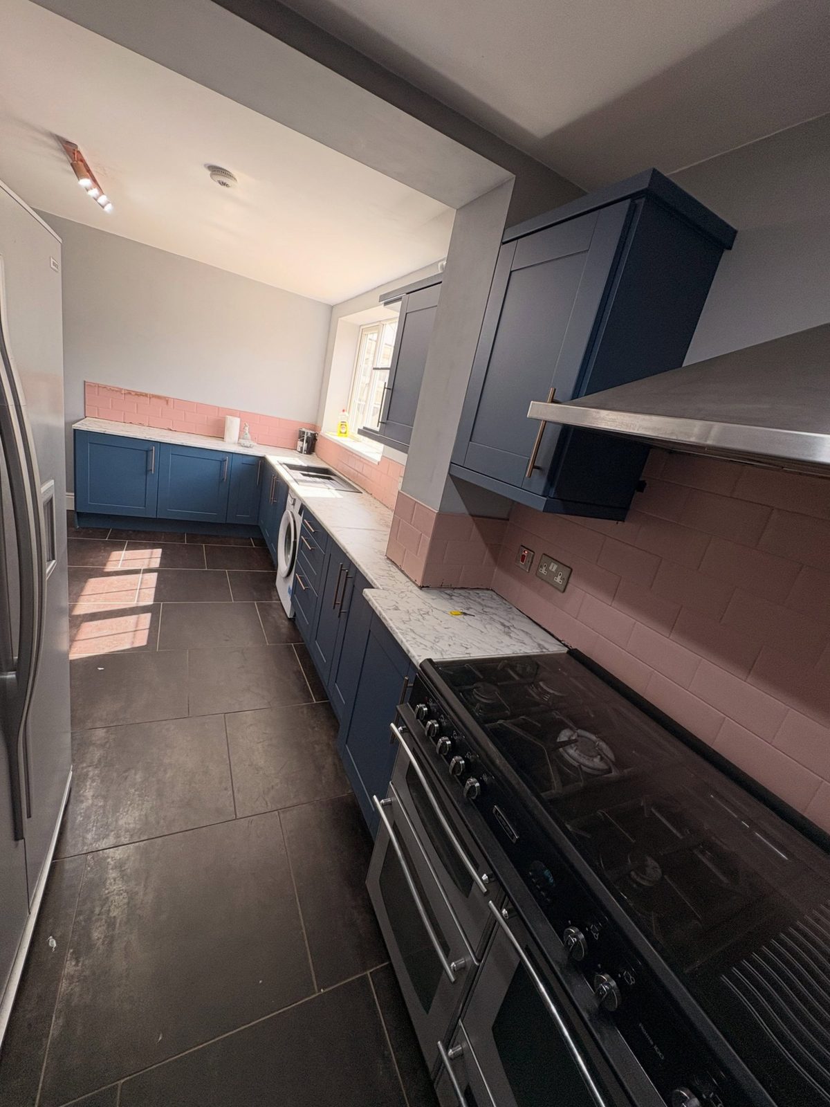 Stiffkey Blue cabinets flanking a black range cooker with stainless hood, pink subway tile behind the hob, marble counter, stainless fridge-freezer on the left, over a dark tile floor.