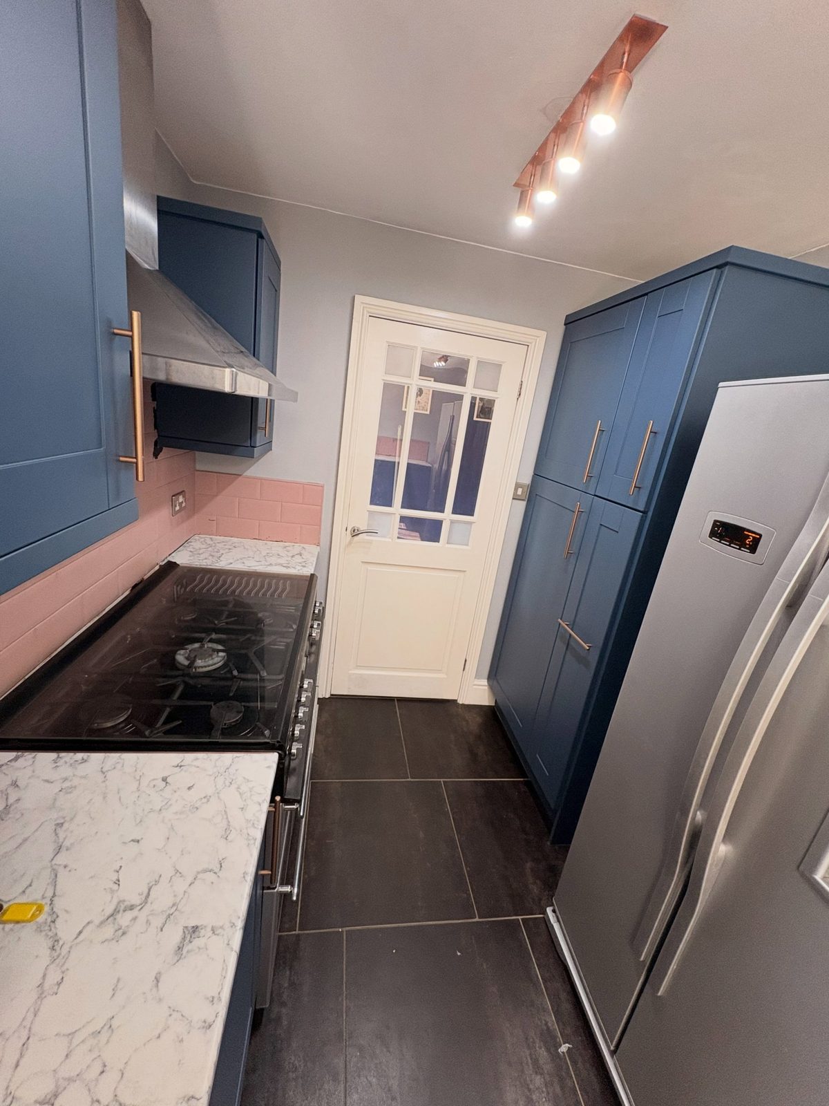 View from the range side showing Stiffkey Blue tall cabinets with copper handles beside a stainless American fridge-freezer, a white glazed door, a copper spotlight track overhead, and a black gas range cooker in the foreground.