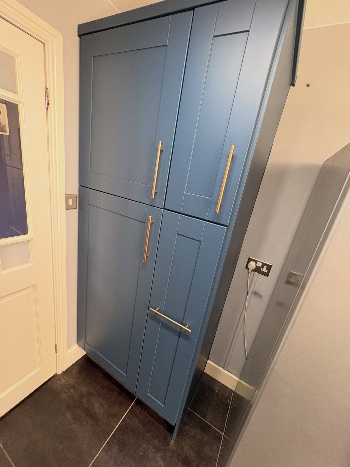 Tall Stiffkey Blue shaker pantry cabinets with long copper bar handles beside a stainless American fridge-freezer, over dark tile floor.