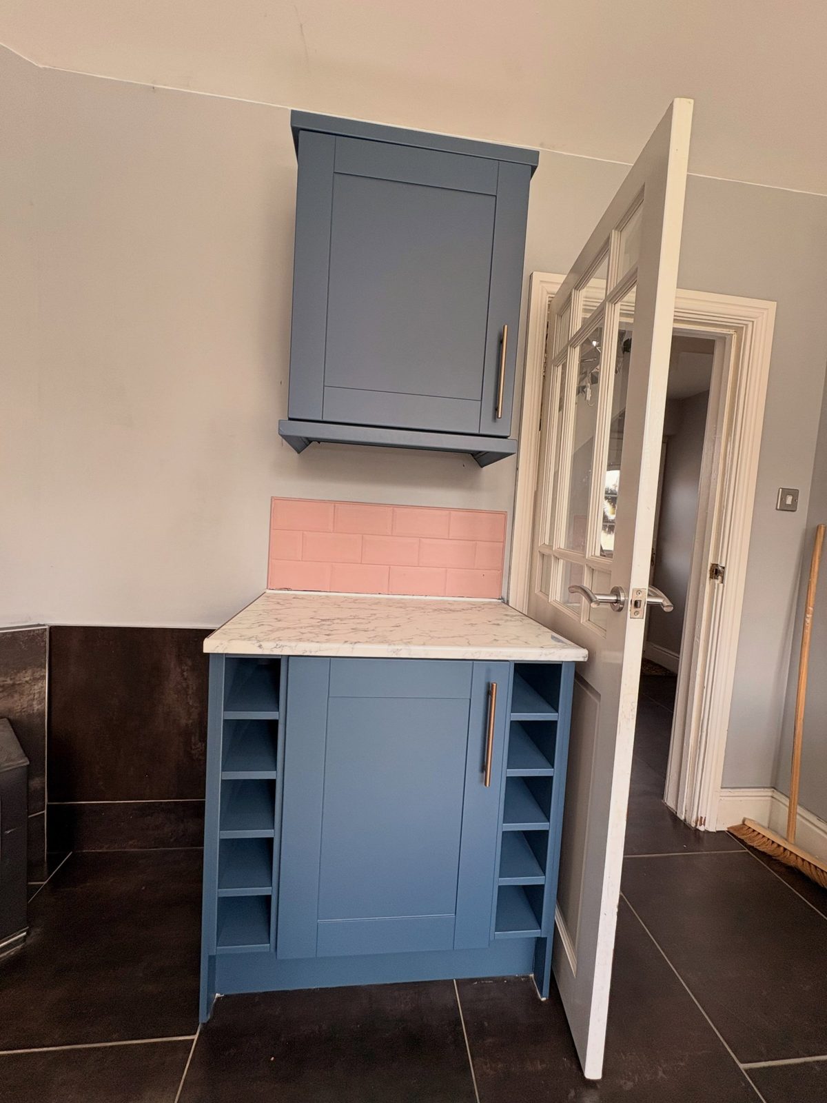 Short Stiffkey Blue base cabinet with built-in wine rack cubbies on either side, pink tile splashback behind, and a small matching wall cabinet above, over dark tile floor.