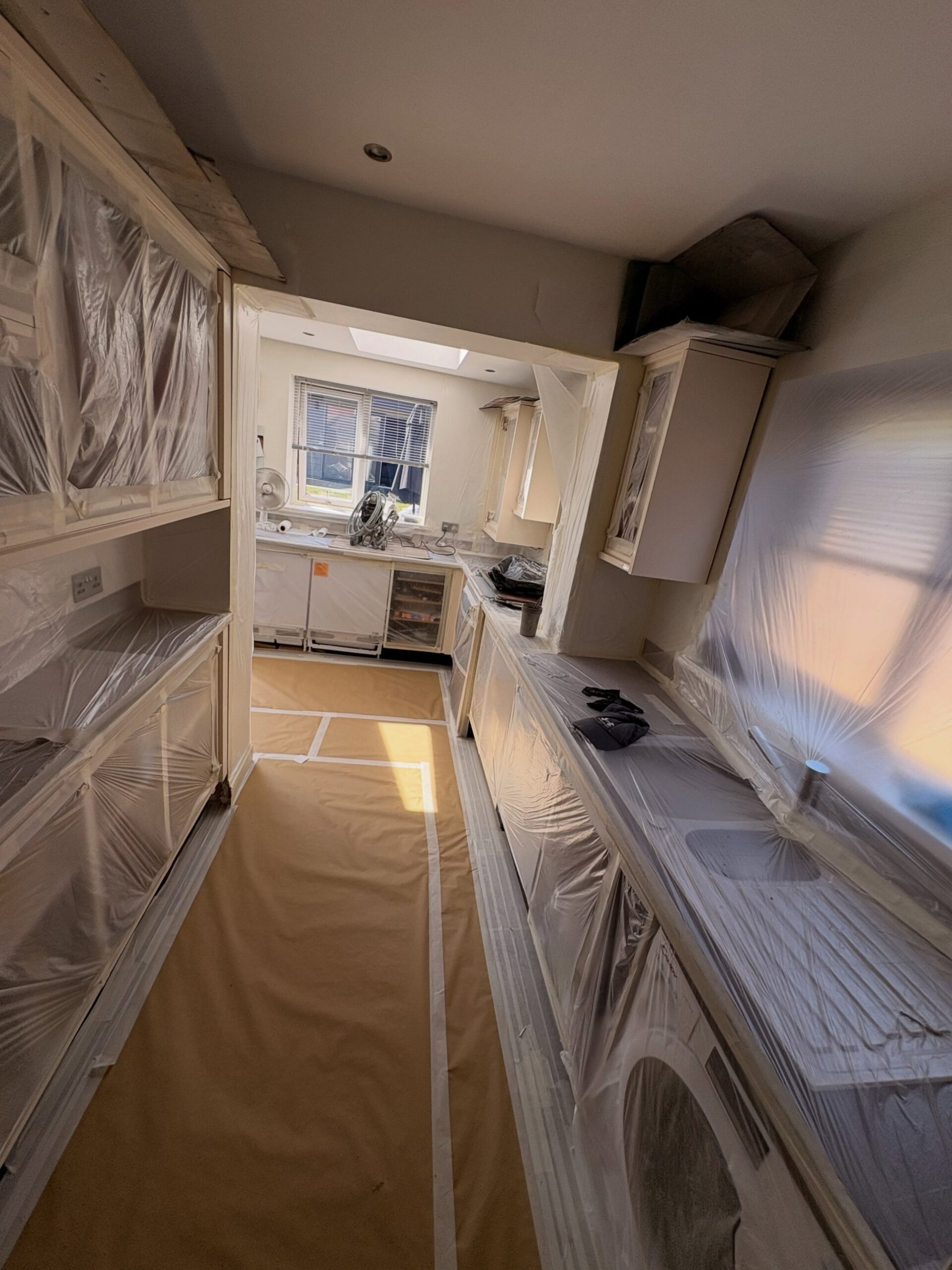 View through a doorway into a kitchen prepped for spraying, cabinets and counters wrapped in plastic, paper runner down the floor, extraction fan running by the far window.