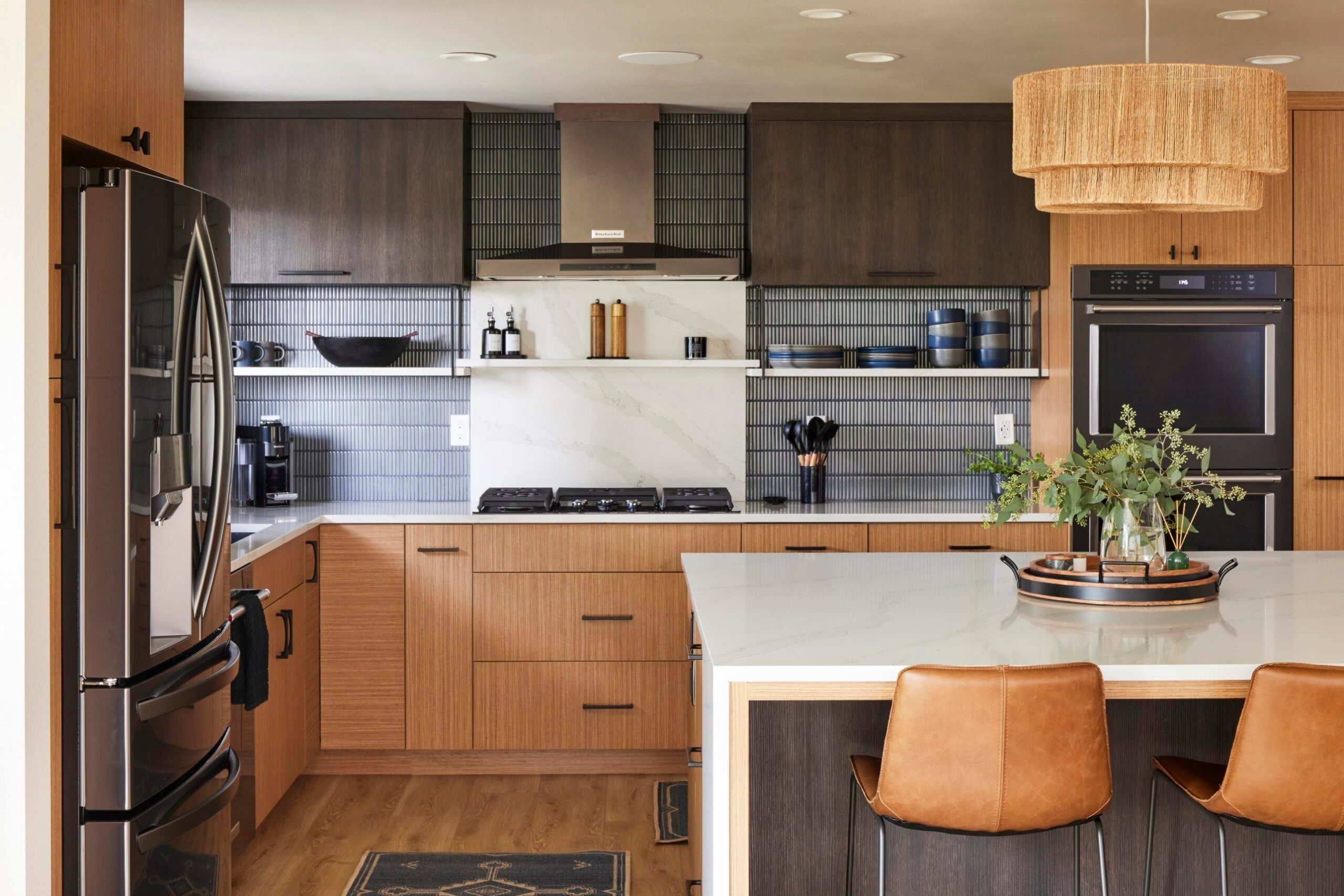 Interior view of the kitchen showing a tiered rattan drum pendant hanging over the island, dark blue vertical tile, open metal cage shelving, a wall oven, a gas cooktop with a stainless hood, and warm teak lower cabinets with dark espresso uppers.