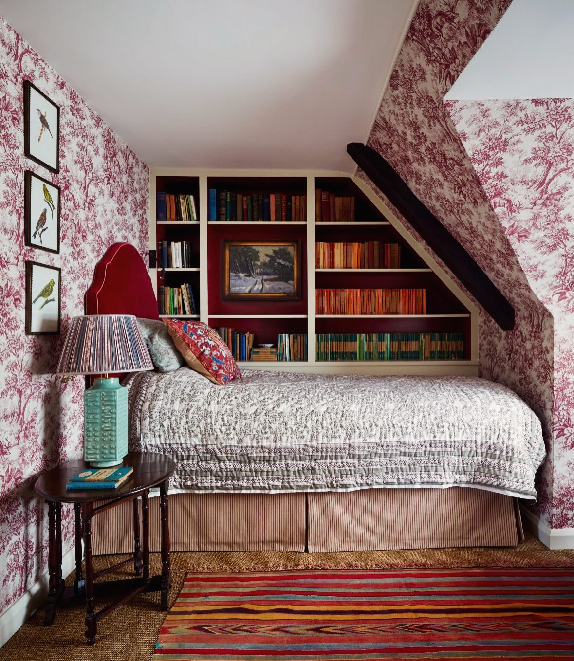 Attic bedroom with pink and white toile wallpaper, built-in bookshelf painted deep red fitted into the sloped wall, red upholstered headboard, and a striped kilim rug