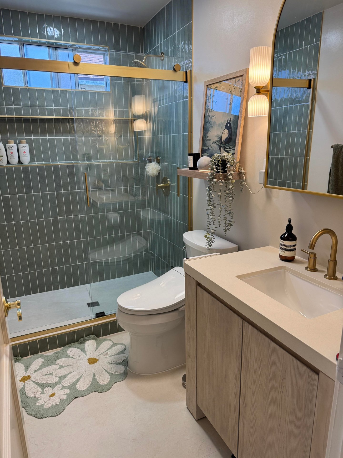 Walk-in shower with sage green vertical stacked tile, frameless glass door, brushed brass hardware, and a daisy-shaped bath mat on the floor outside.