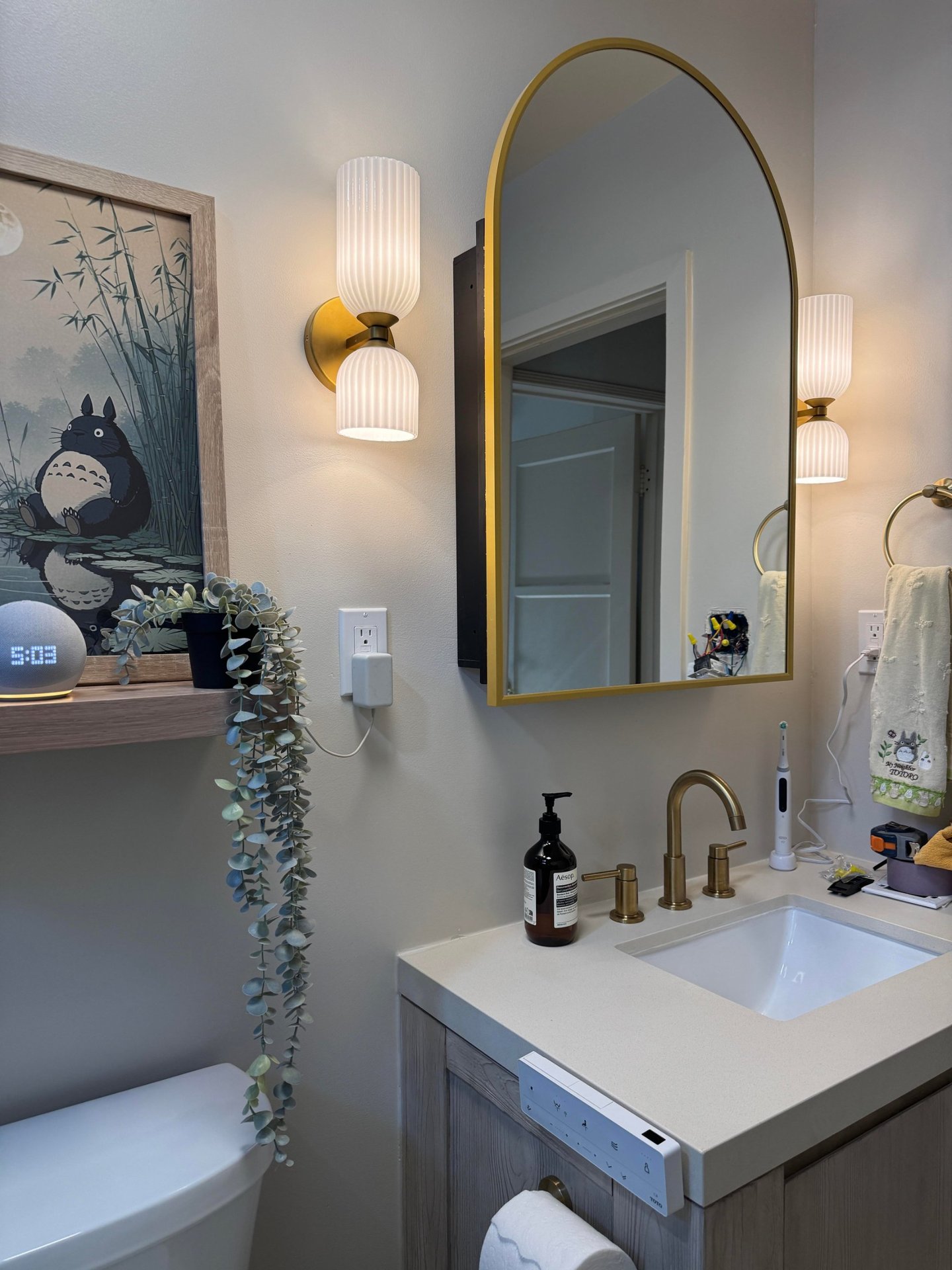Bathroom vanity with a light oak cabinet, white stone counter, undermount sink, arched gold mirror, brass bridge faucet, and an amber Aesop hand wash pump.