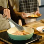 Two people in striped and checkered aprons cooking a breakfast of bacon and sliced sausage in a pastel green nonstick pan on a portable induction cooktop set on a butcher block wood counter, with one hand lifting a piece using stainless steel tongs and the other person holding a plated serving nearby.