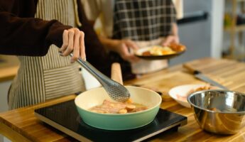 Two people in striped and checkered aprons cooking a breakfast of bacon and sliced sausage in a pastel green nonstick pan on a portable induction cooktop set on a butcher block wood counter, with one hand lifting a piece using stainless steel tongs and the other person holding a plated serving nearby.