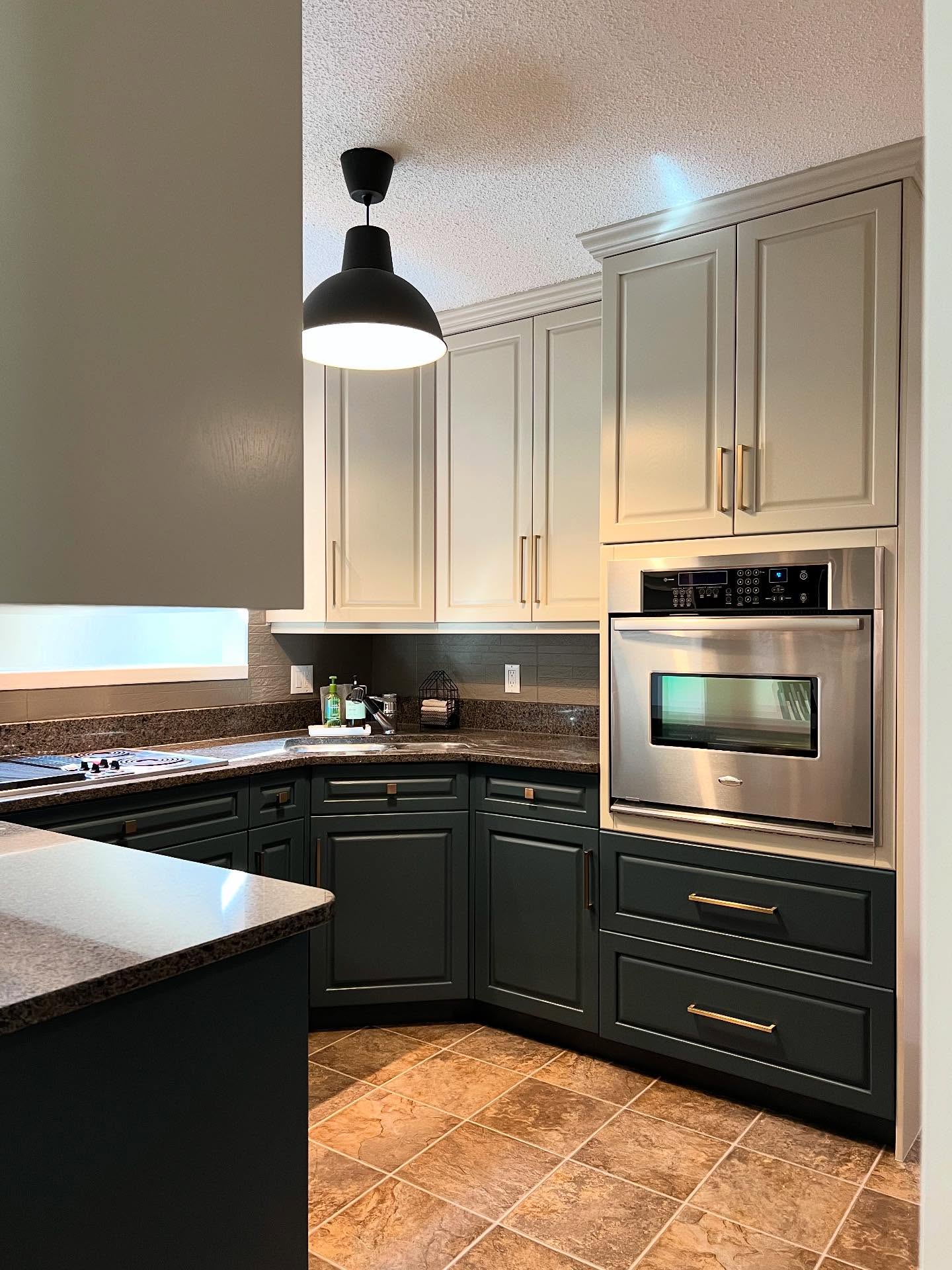 Painted two-tone kitchen corner showing a black electric cooktop, corner sink, built-in stainless Whirlpool wall oven, Hazy Skies sage upper cabinets, Lead Grey dark forest green lower cabinets, long brass bar pulls, speckled granite, a painted gray stone-look backsplash, and a black industrial pendant.