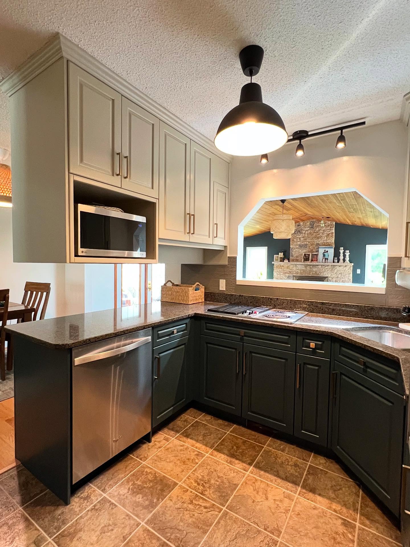 Angled view of the painted two-tone kitchen peninsula with a stainless dishwasher, Lead Grey dark forest green lower cabinets, Hazy Skies sage upper cabinets, LG microwave inset above the counter, speckled granite, and the pass-through window to a stone fireplace living room with a vaulted wood ceiling.