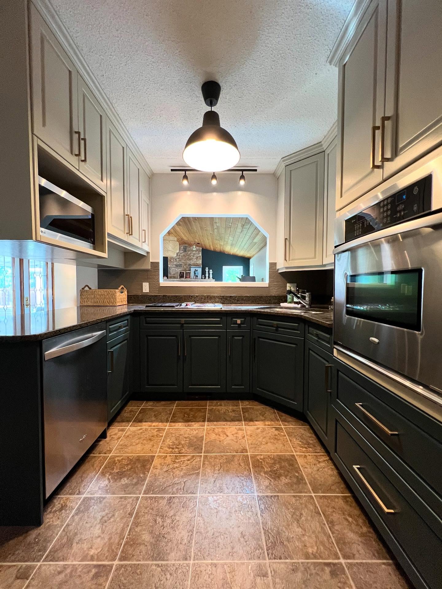 Same U-shaped kitchen with Benjamin Moore Hazy Skies sage upper cabinets, Lead Grey dark forest green lower cabinets, long brass bar pulls, a black industrial pendant with three-head track lighting, dark painted backsplash, and the pass-through window into the stone fireplace living room.