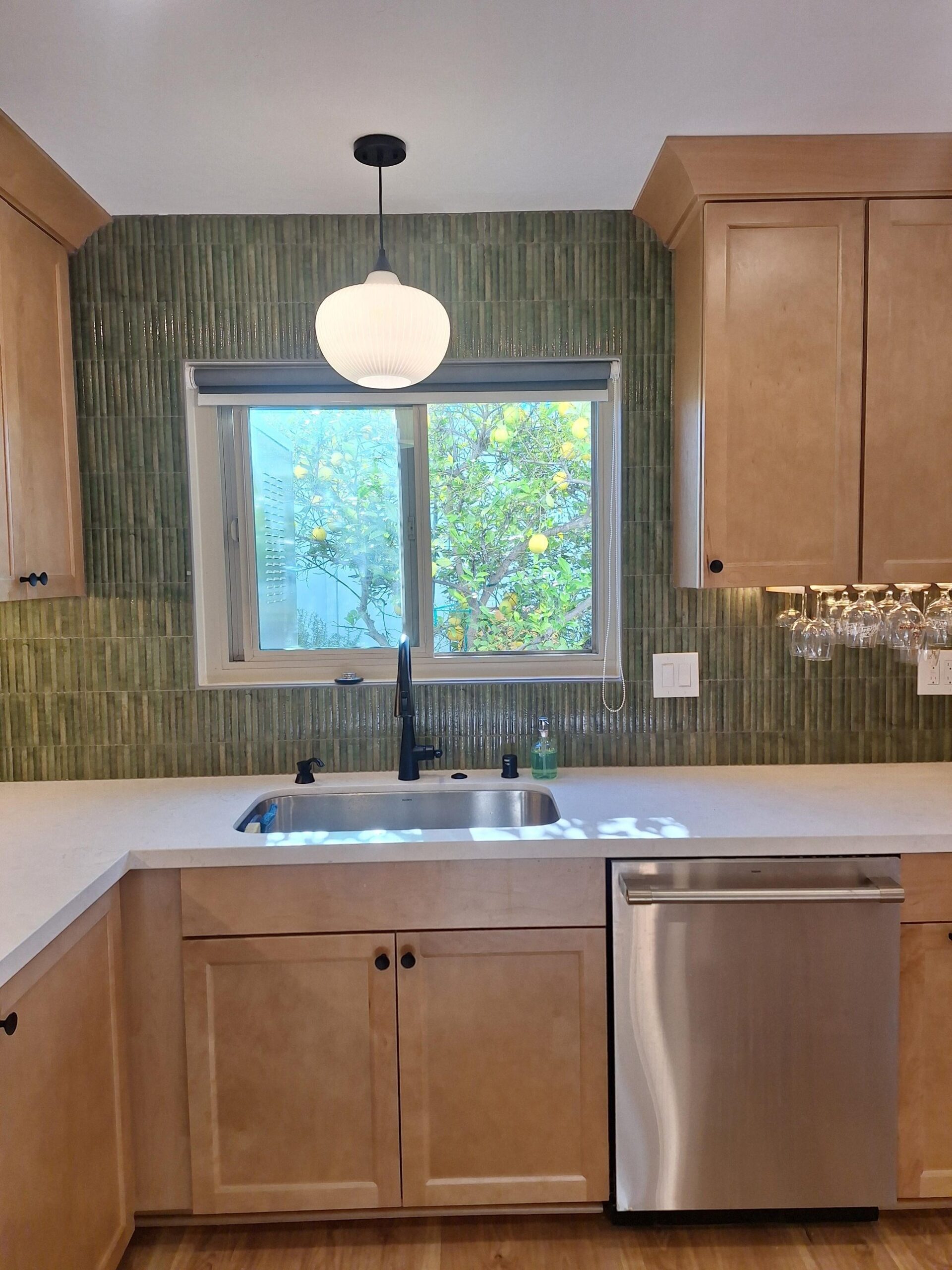 Honed quartz counter with a stainless undermount sink, matte black pull-down faucet, green vertical fluted ceramic tile running floor to ceiling, and a window looking out at a lemon tree.