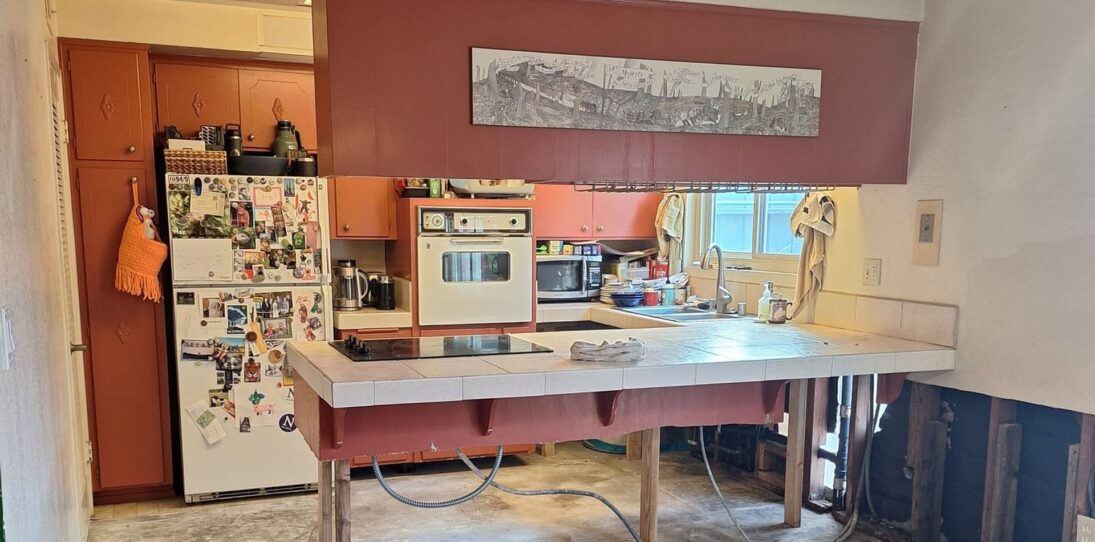 1966 kitchen mid-demolition with deep Venetian red cabinets, a vintage white General Electric wall oven, a smooth cooktop set into a tiled peninsula propped up on raw 2x4 legs, a magnet-covered white top-freezer fridge, flooring stripped down to concrete slab, and an open wall base with studs and plumbing visible.