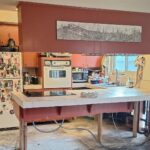 1966 kitchen mid-demolition with deep Venetian red cabinets, a vintage white General Electric wall oven, a smooth cooktop set into a tiled peninsula propped up on raw 2x4 legs, a magnet-covered white top-freezer fridge, flooring stripped down to concrete slab, and an open wall base with studs and plumbing visible.