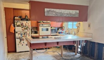 1966 kitchen mid-demolition with deep Venetian red cabinets, a vintage white General Electric wall oven, a smooth cooktop set into a tiled peninsula propped up on raw 2x4 legs, a magnet-covered white top-freezer fridge, flooring stripped down to concrete slab, and an open wall base with studs and plumbing visible.