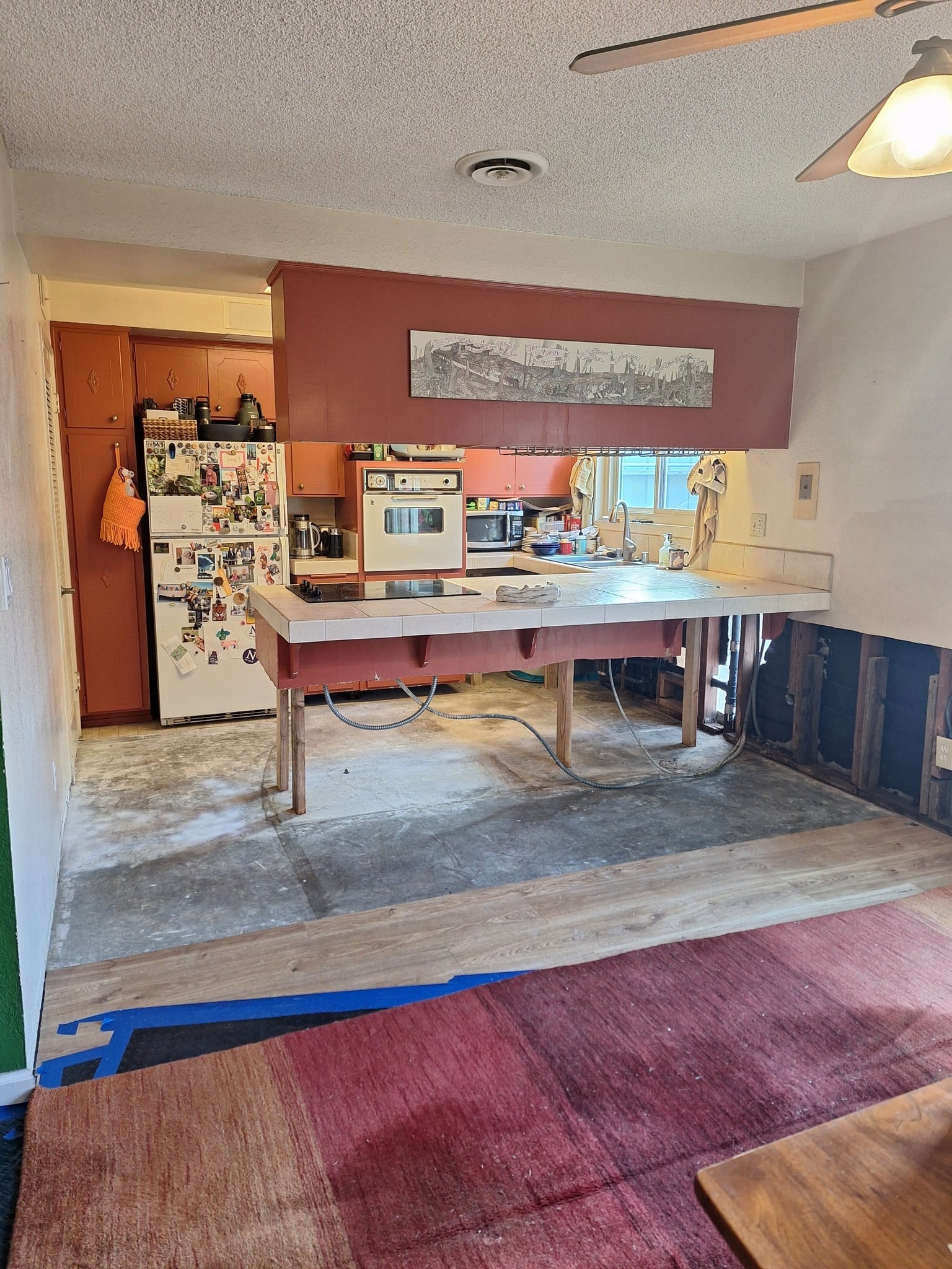 Mid-demolition kitchen with deep Venetian red cabinets, the tiled peninsula propped up on raw 2x4 legs, flooring stripped down to concrete, and an open wall base with studs and plumbing visible.