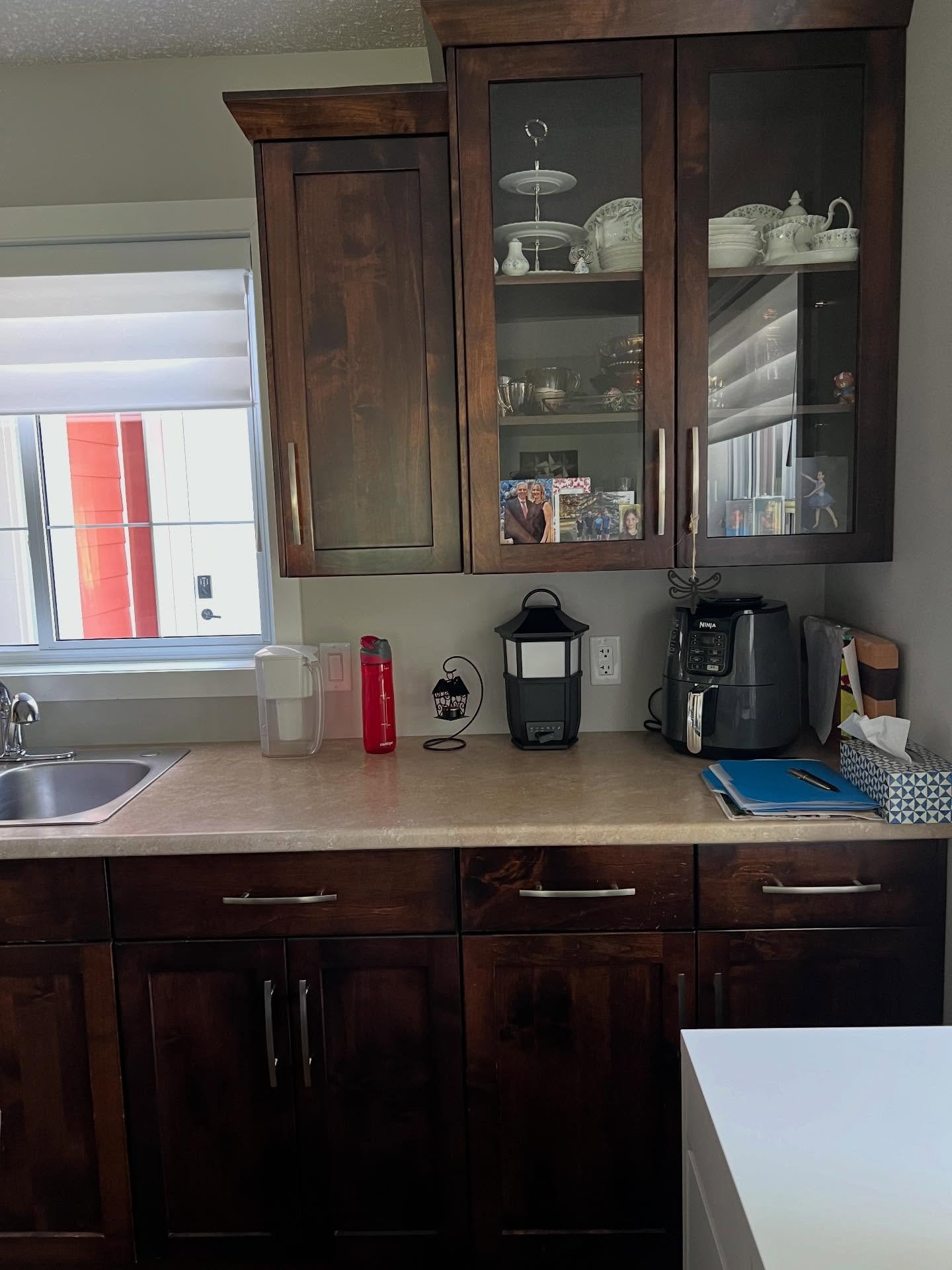 Dark walnut glass-front display hutch filled with white china, a teapot, silver serving pieces, and family photos, next to a window with closed blinds and a red building visible outside, with a bar sink on the counter below.
