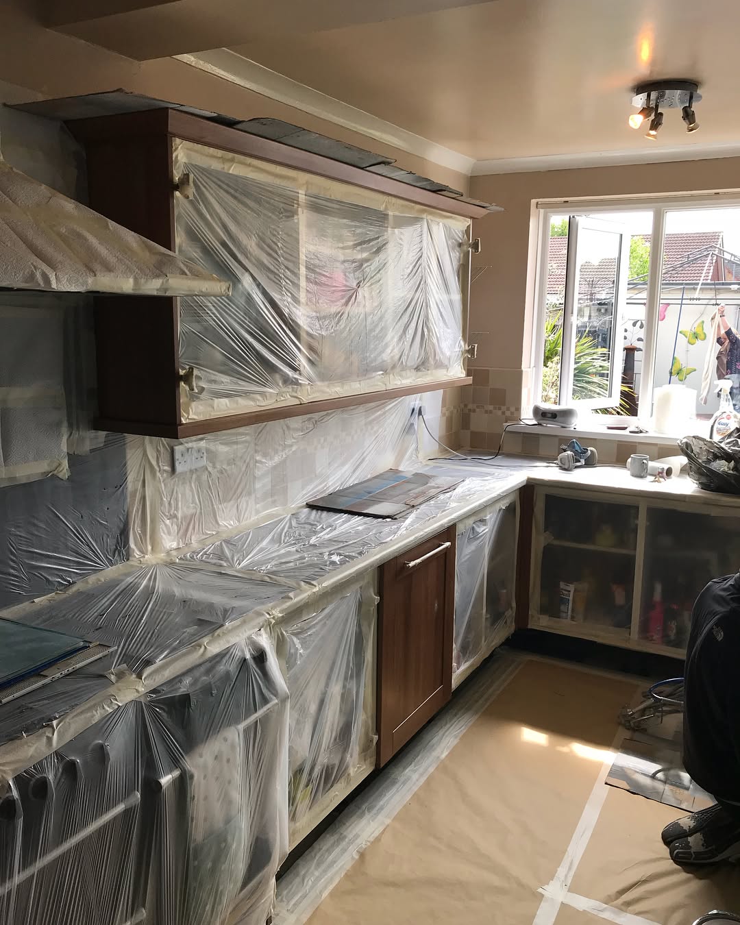 Walnut kitchen cabinets covered in plastic sheeting and masking paper, counters wrapped, floor papered over, with respirator and ladder visible as the sprayer preps the room.