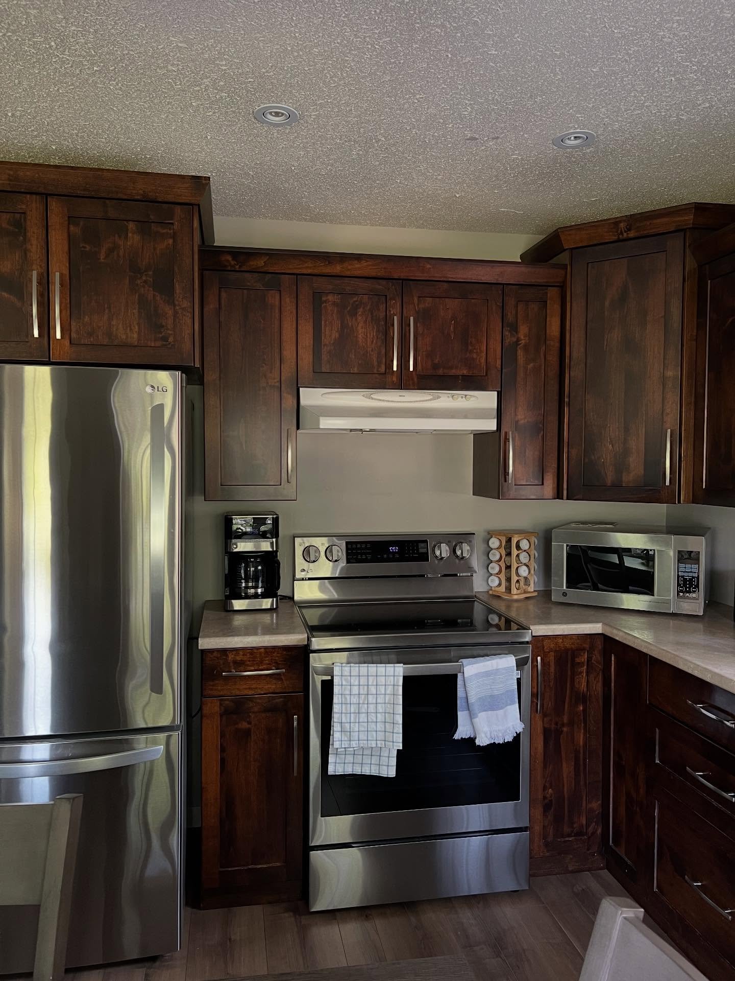 Dark walnut shaker kitchen cabinets around a stainless electric range with a white range hood, LG stainless fridge on the left, drip coffee maker on the counter, tan speckled laminate counters, and dark wood vinyl plank flooring.