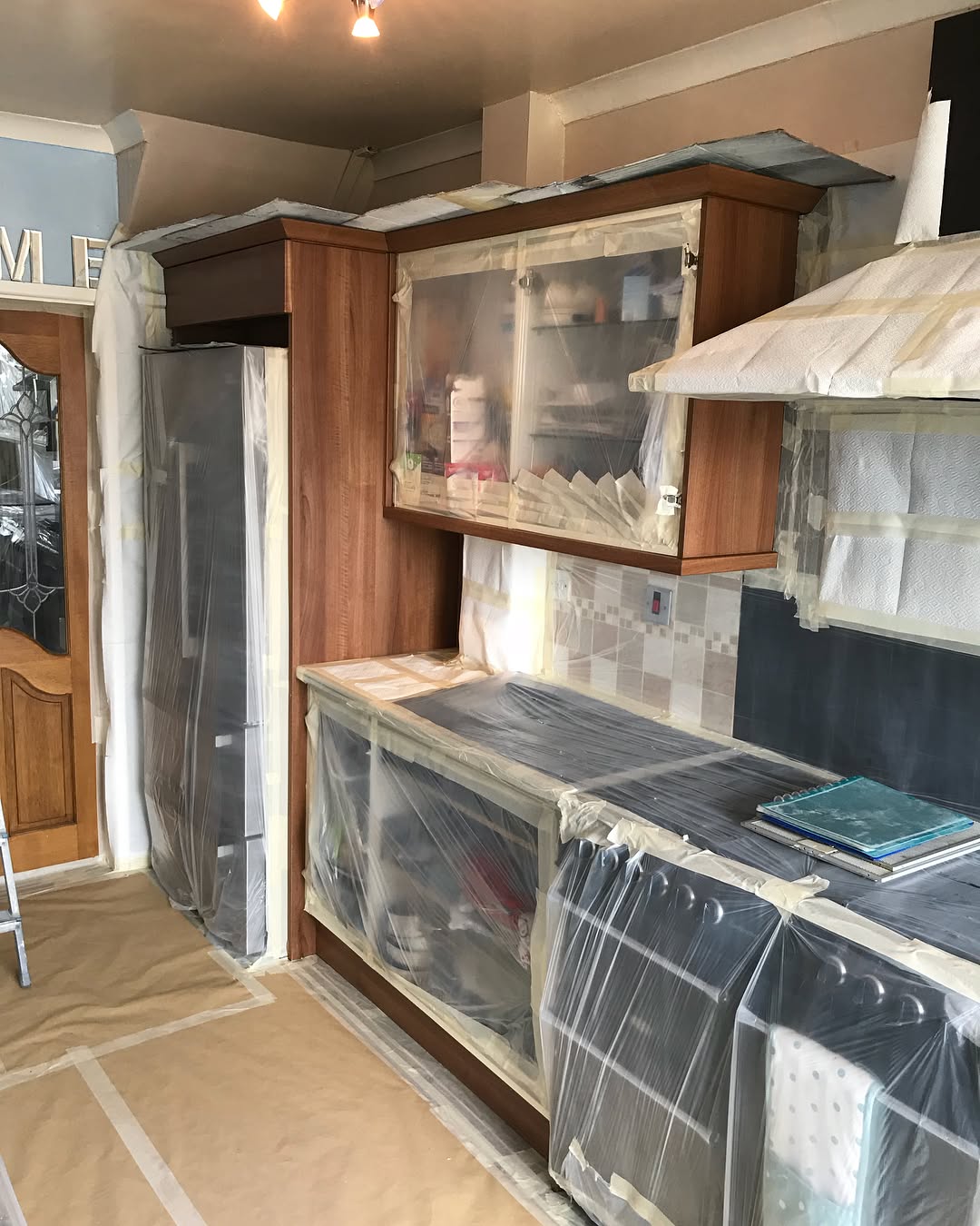 A closer view of the same prep stage with walnut cabinet frames still visible, extractor hood masked with paper towel, fridge wrapped in clear plastic, and the backsplash taped off.