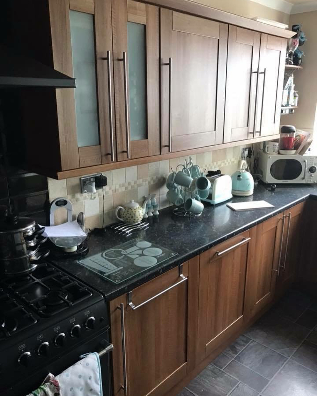 Wide view of a walnut Shaker kitchen with glossy black granite counters, a gas range on the left, pastel teal kettle and toaster, mugs on a stand, and warm wood tones throughout.