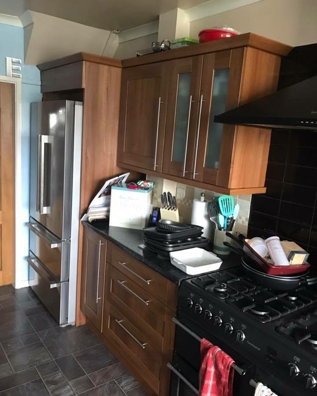 Walnut kitchen with a tall American style fridge freezer on the left and a black range cooker on the right, with pans and bakeware stacked on the counter and dark tile floor.
