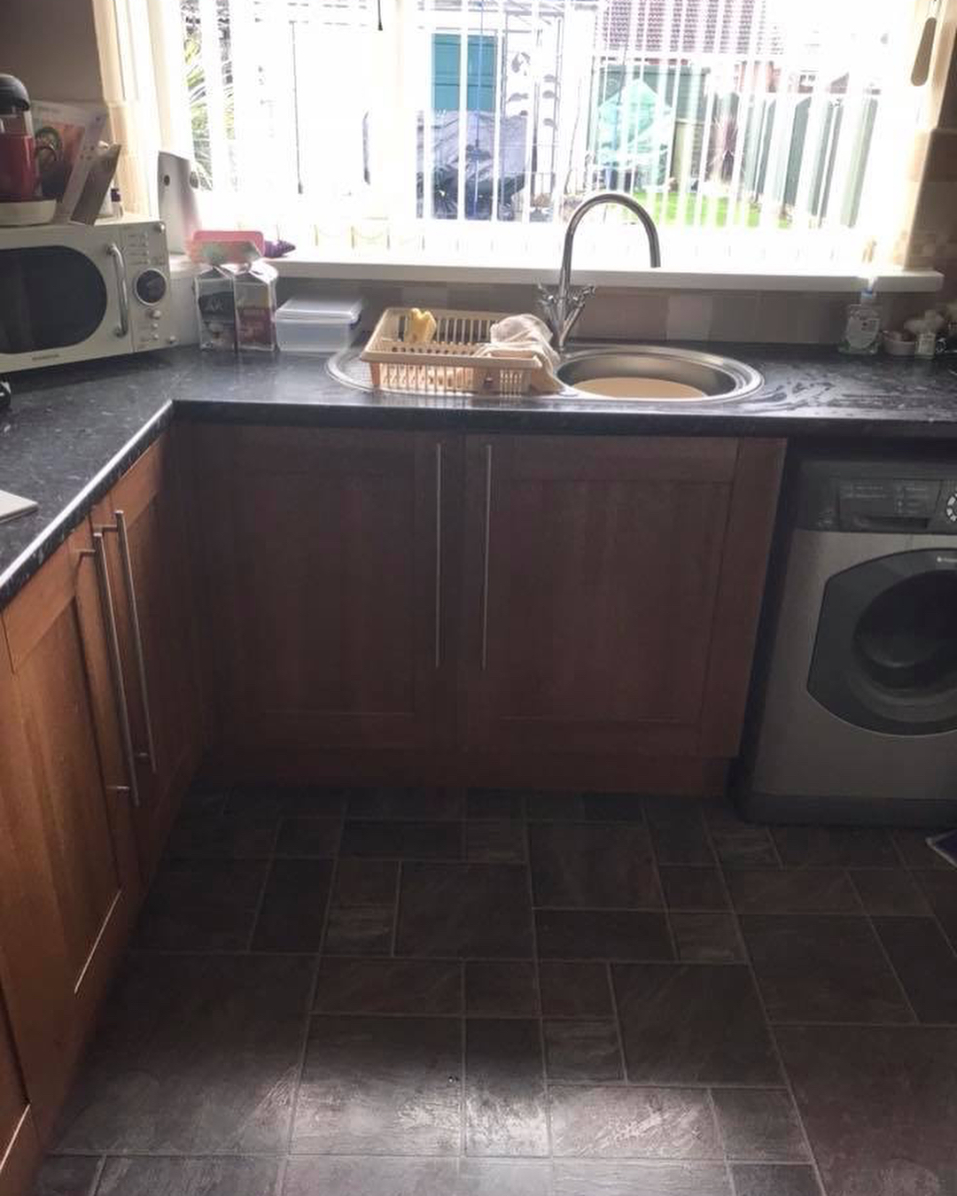 Walnut kitchen corner with the sink under a bright window, dish rack on the counter, washing machine beside the base cabinets, and dark slate style tile on the floor.