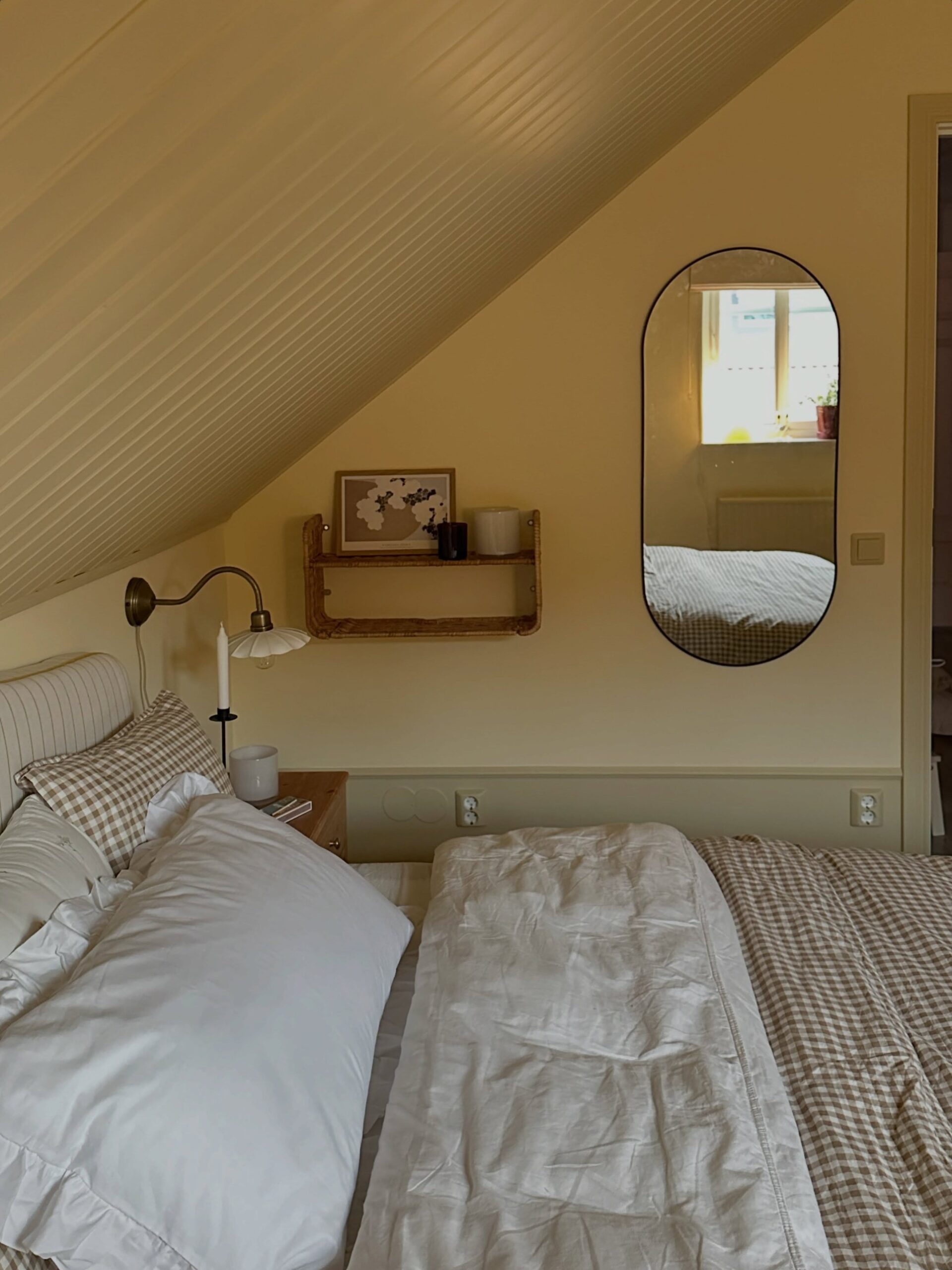 Attic bedroom at golden hour with warm yellow walls, ribbed vertical wood ceiling painted white, gingham brown and cream bedding, oval mirror, rattan wall shelf, and a lit candle on the nightstand