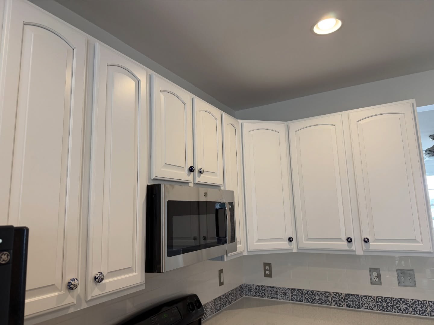 Low-angle view looking up at white arched raised-panel upper cabinets, with blue and white ceramic knobs, a stainless microwave below, a recessed ceiling light, and the Moroccan tile backsplash visible at the bottom.