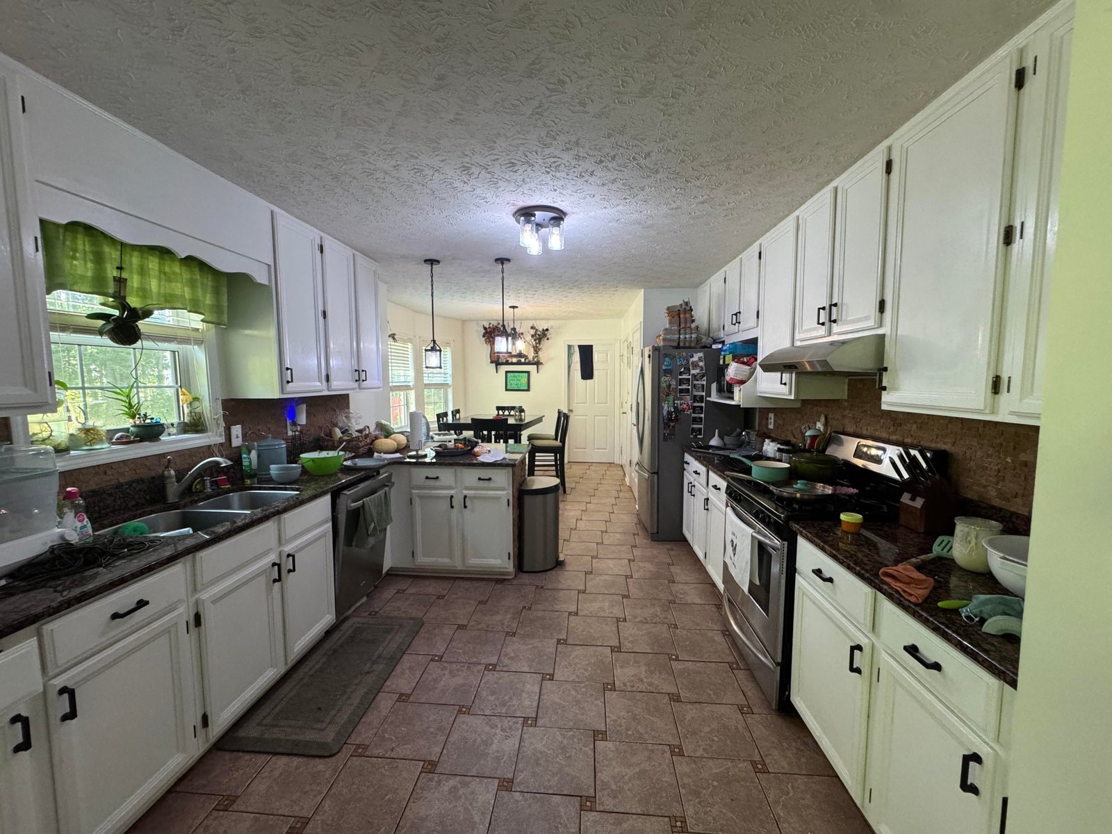 A long galley kitchen with white cabinets, a brown cork-look tile backsplash, speckled dark granite countertops, a brown basket-weave tile floor, a scalloped green valance curtain over the sink window, and cluttered countertops.