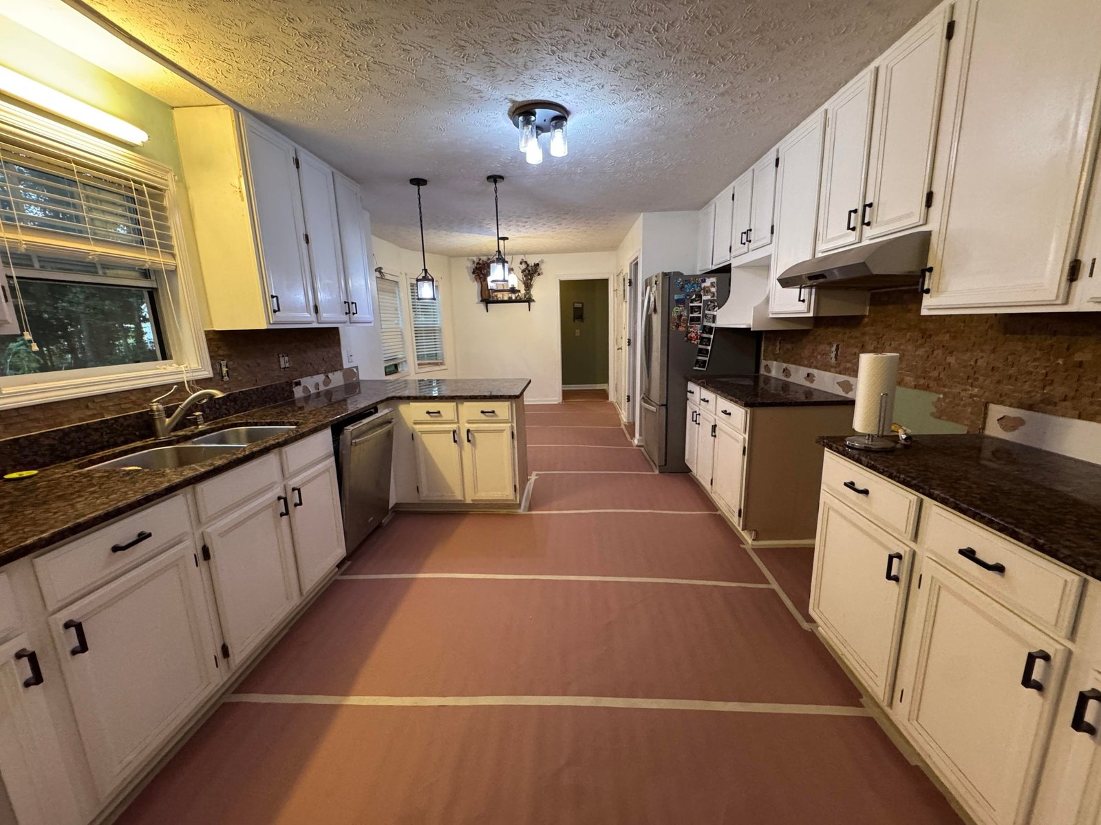 The same kitchen mid-renovation with the old backsplash torn off, the floor covered in overlapping orange construction paper taped at the seams for protection, white cabinets still in place, and empty countertop runs.