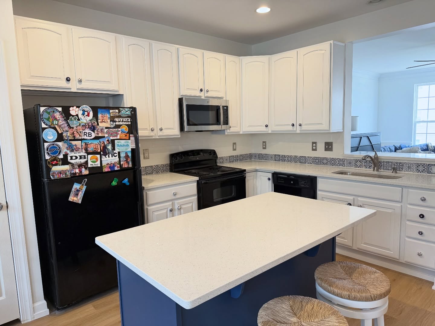 Kitchen with Sherwin-Williams Pure White cabinets on the perimeter, a deep blue Indigo Batik island with a white quartz top, a black fridge, a black stove, a black dishwasher, white speckled quartz counters, two rush-seat stools, and the original Moroccan tile backsplash tying the whole room together.